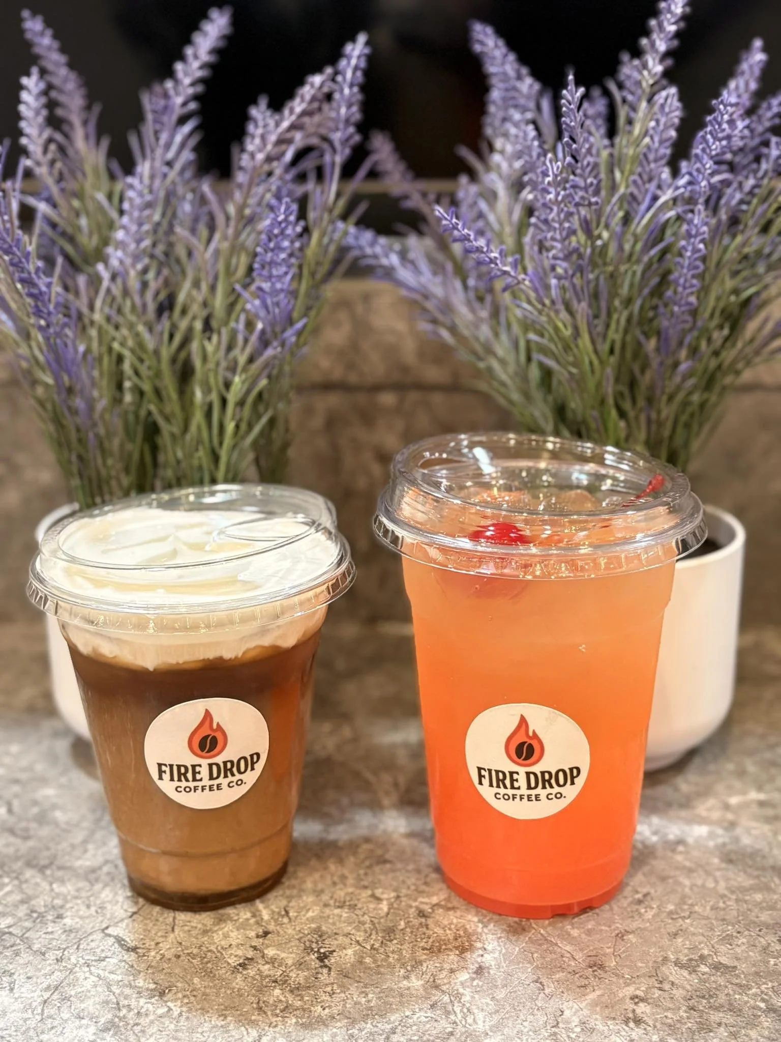 Two plastic cups with Fire Drop Coffee Co. logos on a table, one containing a cold brew coffee with cream and the other a pinkish fruit drink, with potted lavender plants in the background.
