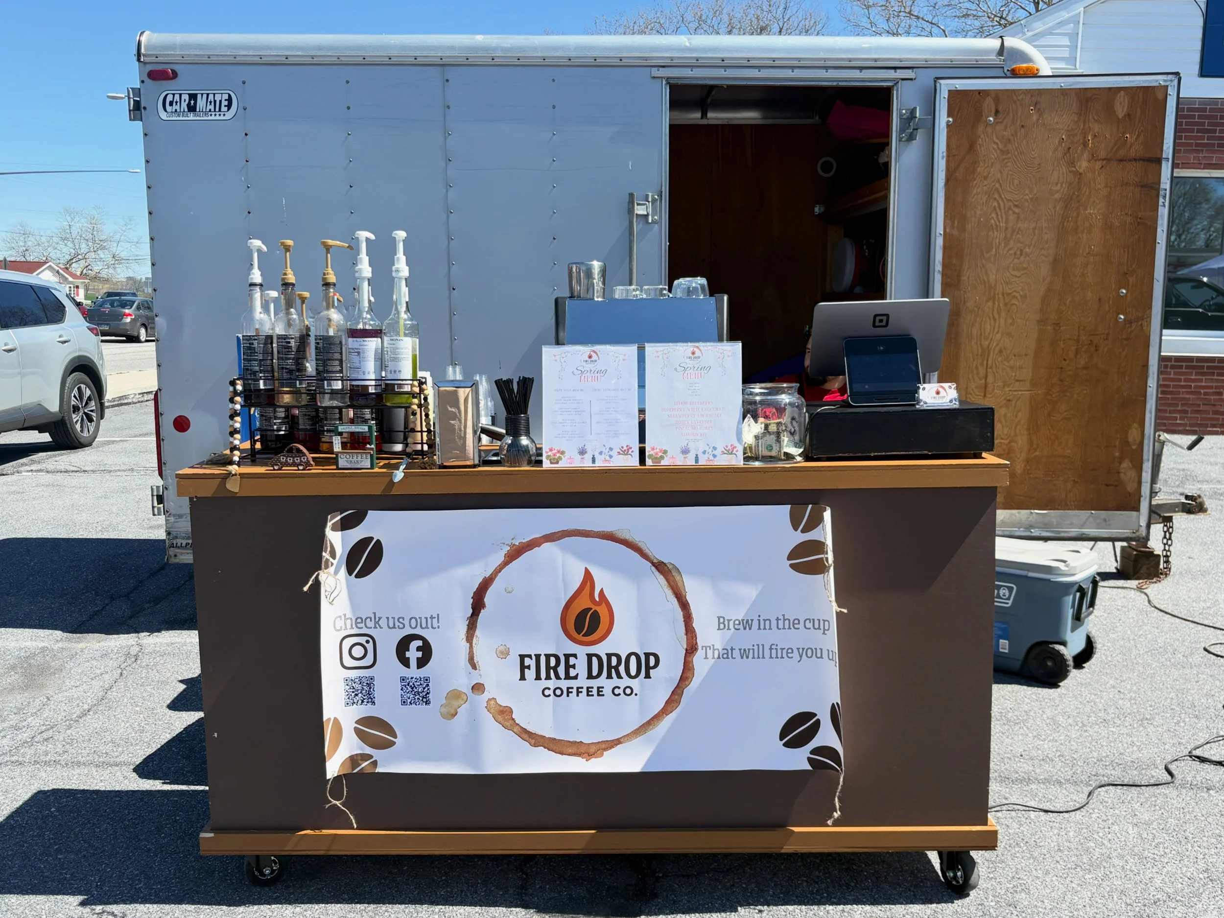 A coffee stand named Fire Drop Coffee Co. with bottles of flavored syrups, a menu, and a cash register, set up outdoors on pavement with cars in the background.