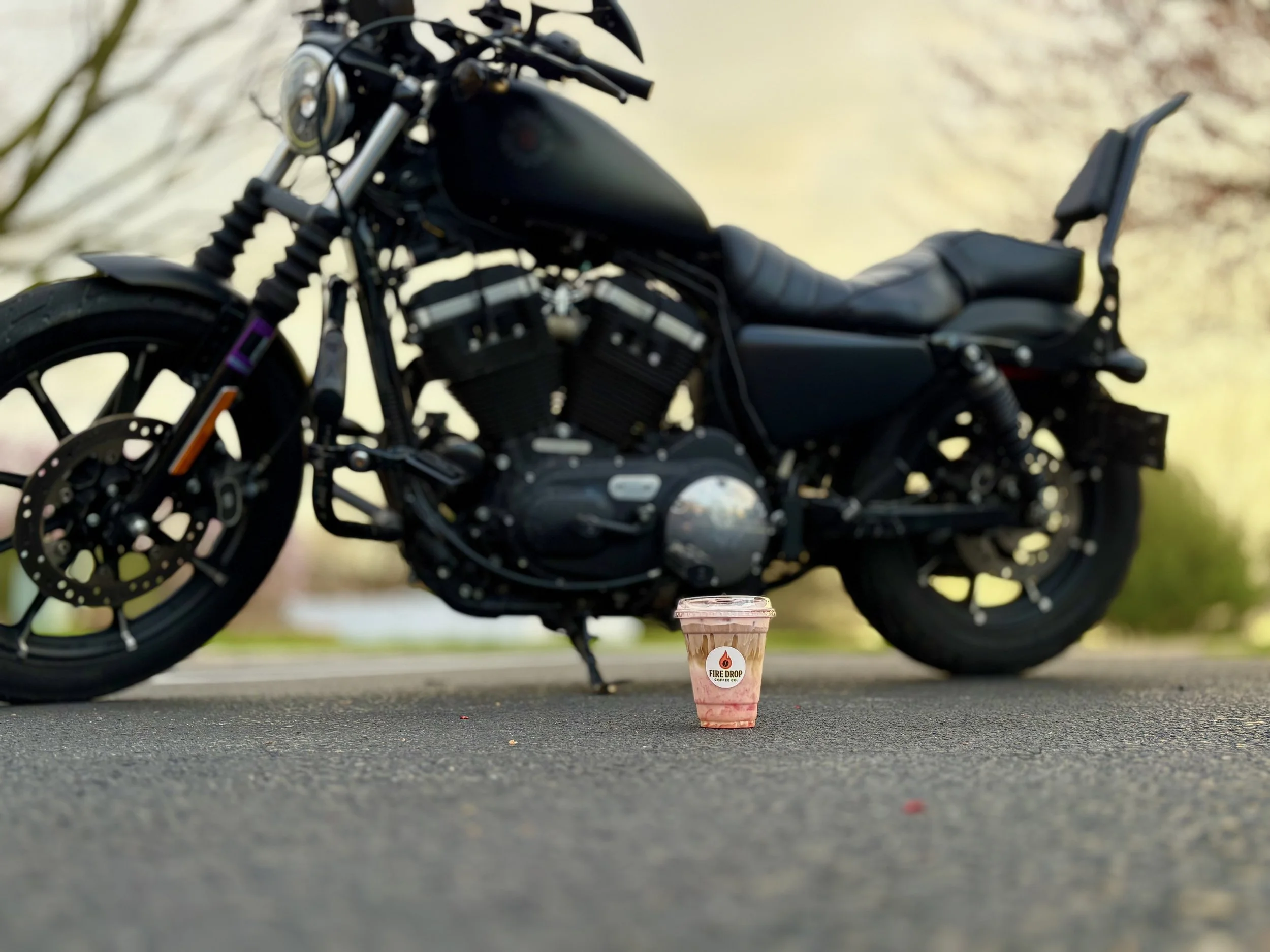 Black motorcycle parked on pavement with a small pink and white frappuccino in front, labeled 'Fire Drop' coffee, during daytime with blurred background.