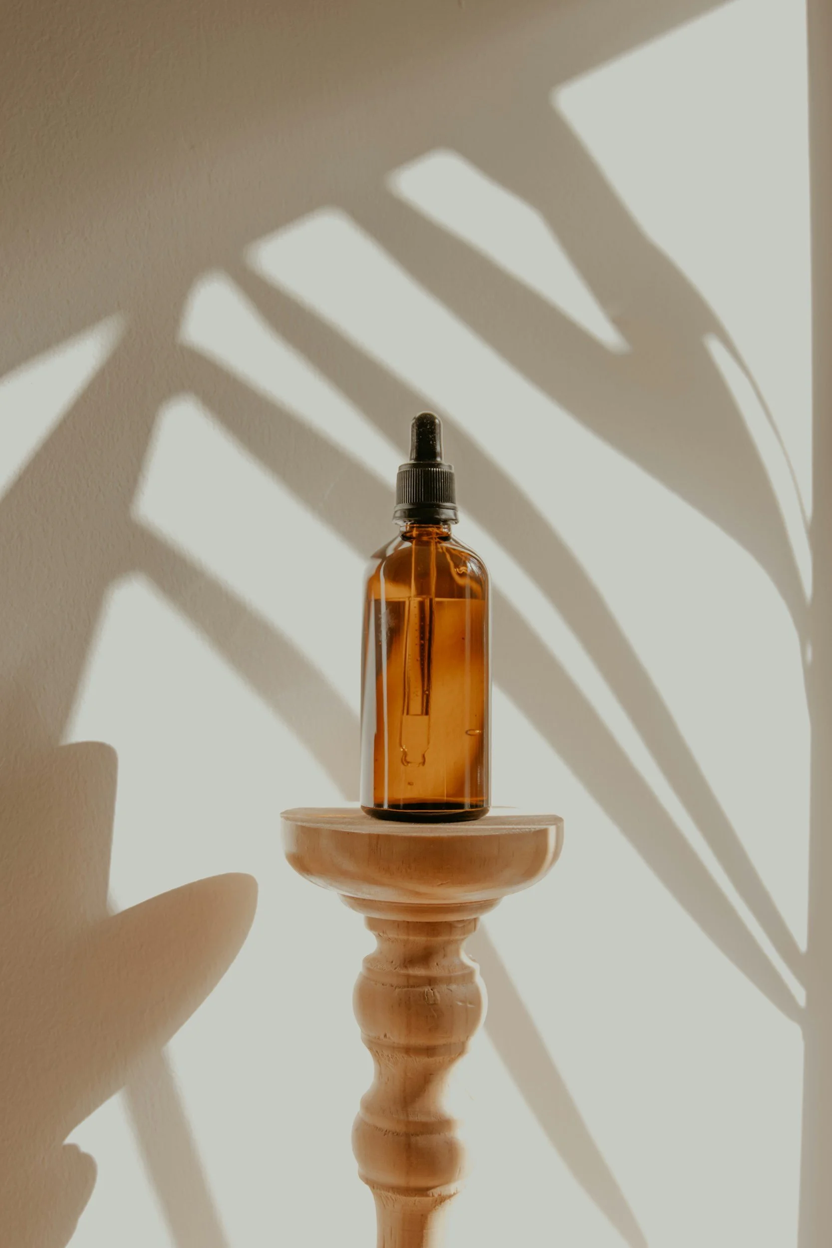 A brown glass dropper bottle with a black cap sitting on a wooden pedestal, with shadows of large leaves cast on the wall behind.