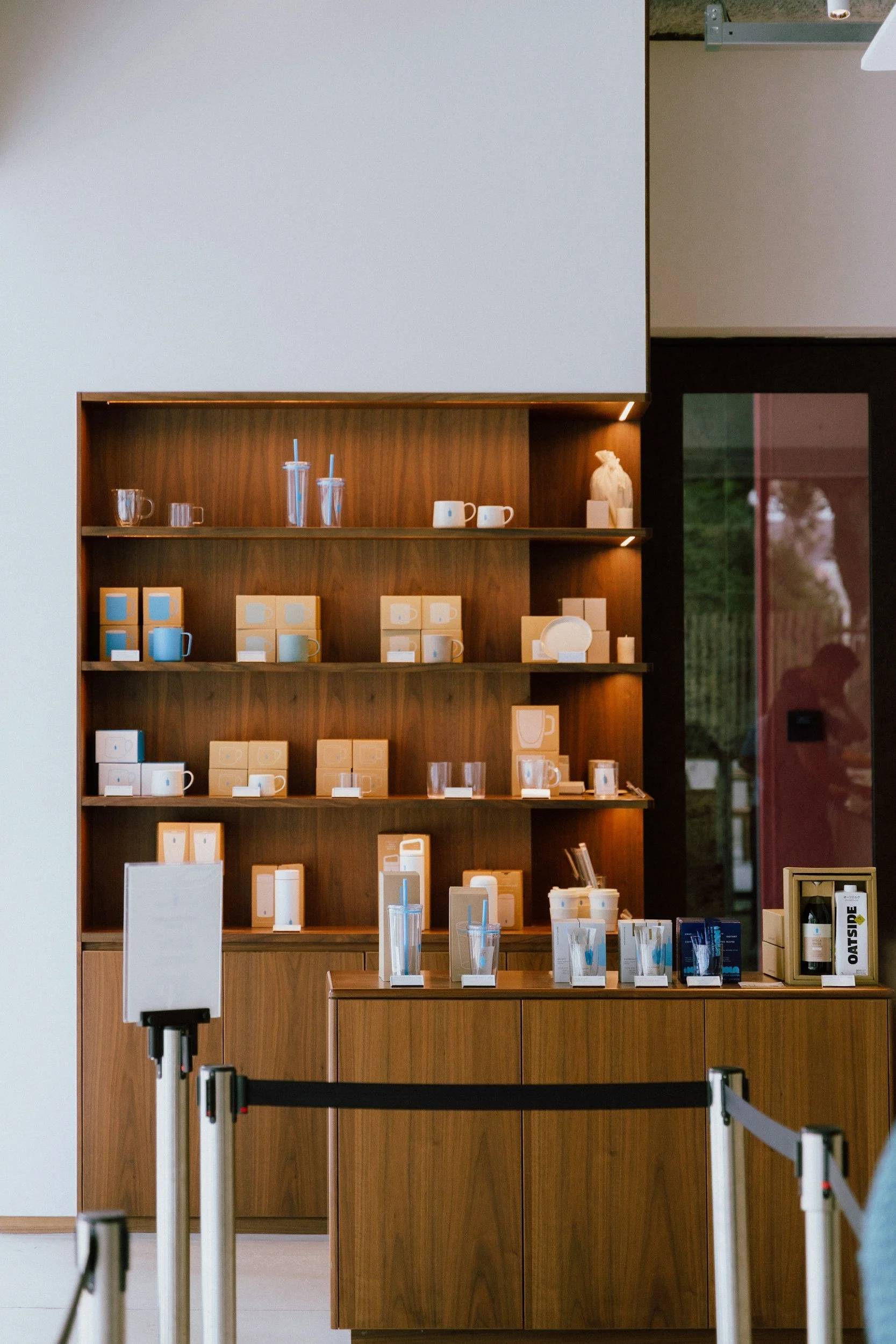 Indoor display of glassware, cups, and packaging on wooden shelves in a store, with a glass door and people reflected in it.