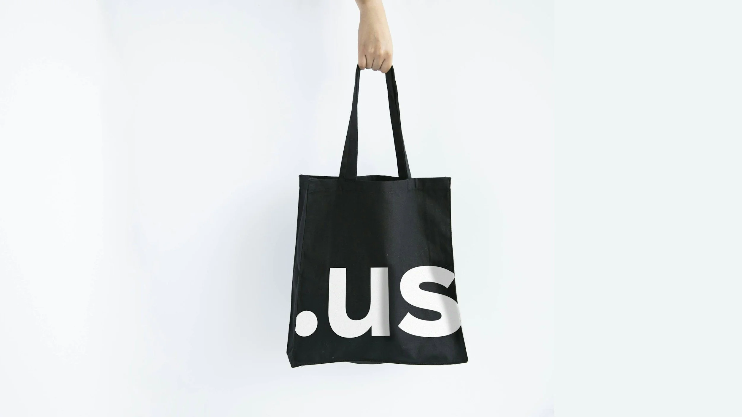 A black tote bag with the partial text '.us' printed in white, held by a person's hand against a plain white background.