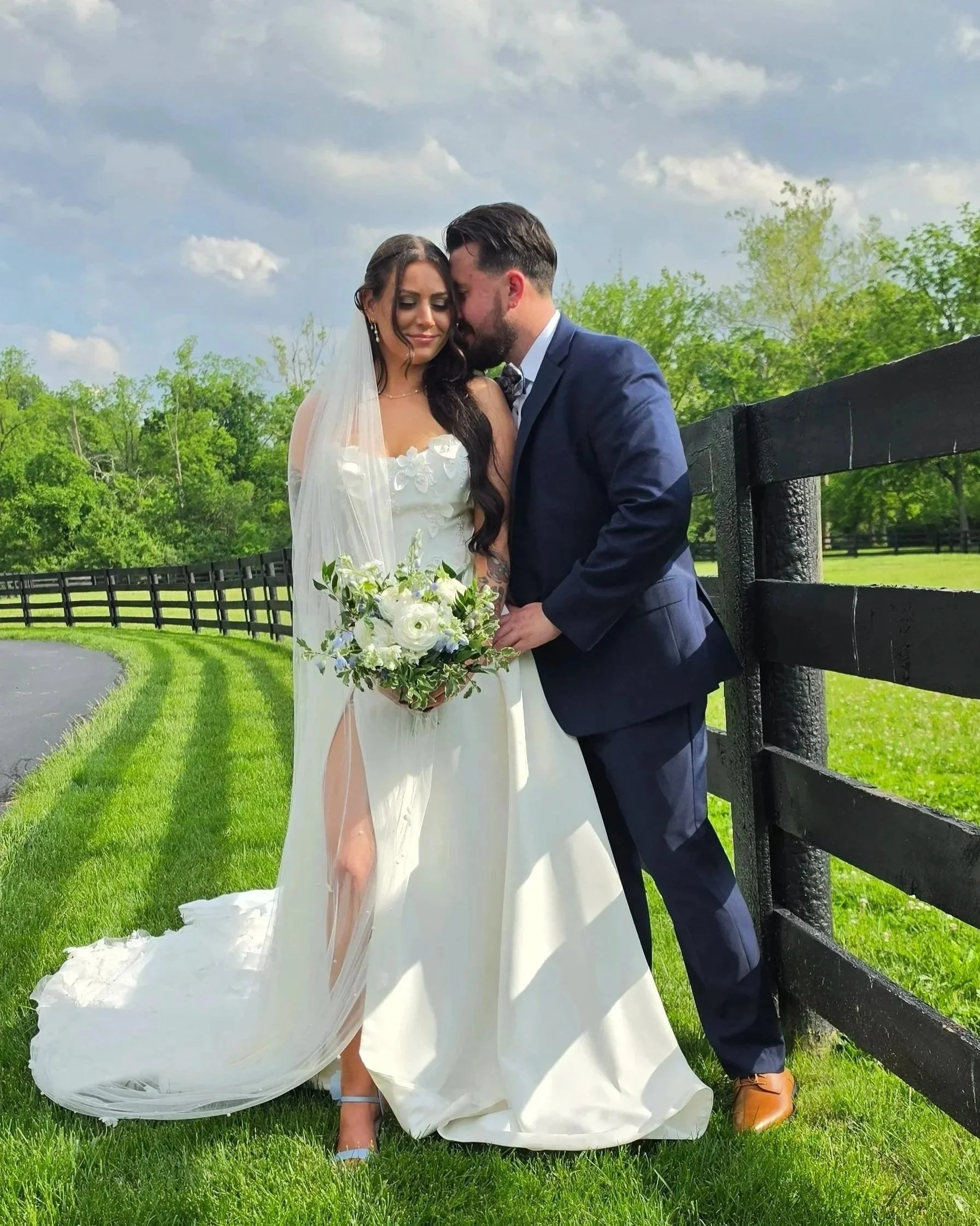 A bride and groom standing close together outdoors near a black wooden fence, with green trees and a cloudy sky in the background. The bride is wearing a white wedding dress with detailed lace and holding a bouquet of white flowers. The groom is wearing a navy blue suit and brown shoes. They are sharing an intimate moment, with their foreheads touching, eyes closed, and smiling gently.