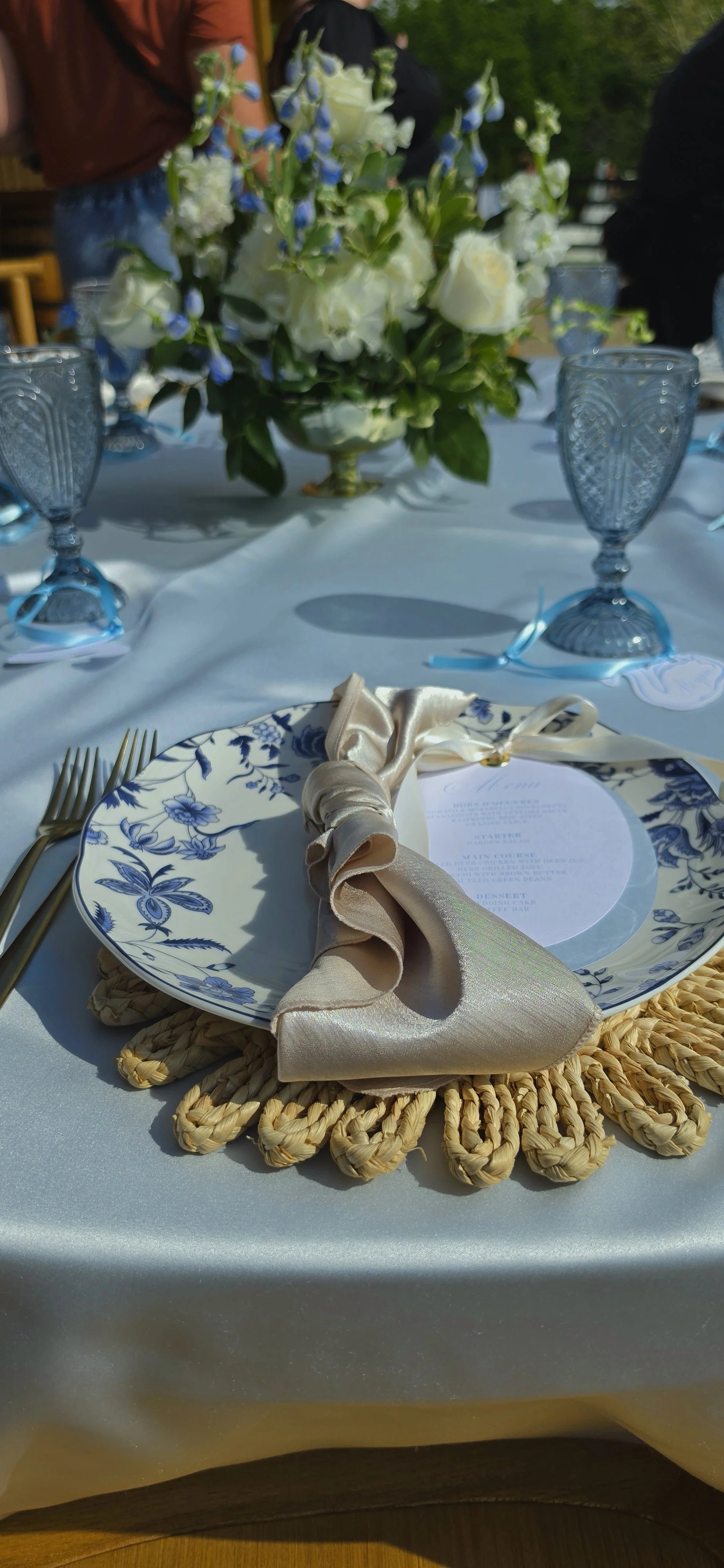 A table setting with a white and blue floral plate, a beige napkin tied with a ribbon, gold utensils, and a woven placemat. In the background, there is a floral centerpiece with white roses and blue flowers. Several blue glass goblets are arranged around the table.