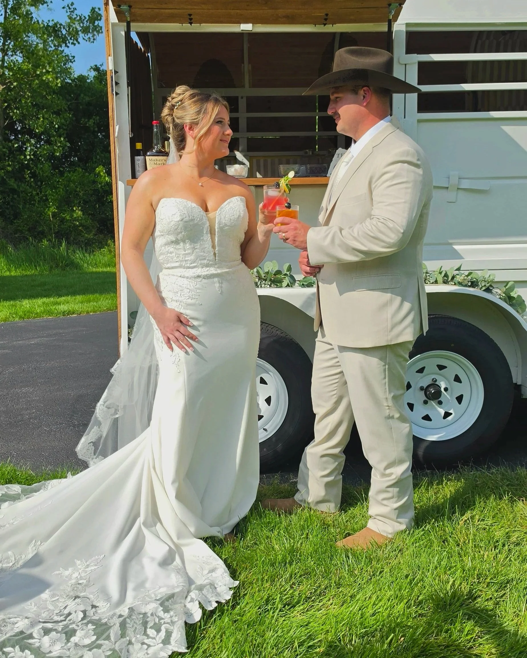 A bride and groom standing in front of a trailer during their outdoor wedding, holding drinks and looking at each other with trees and grass in the background.