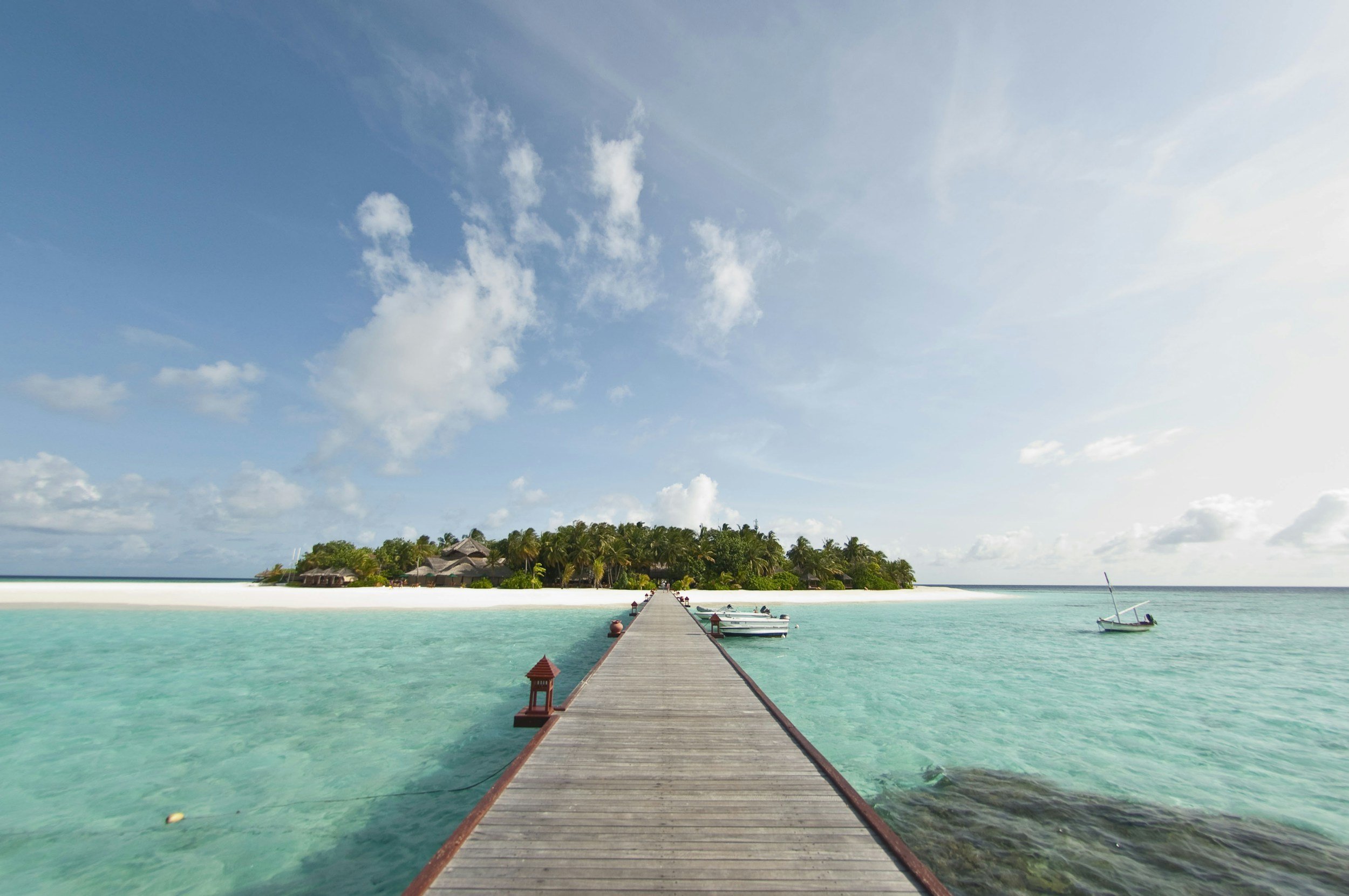 A wooden pier extending over clear turquoise water towards a tropical island with palm trees and thatched-roof buildings, under a partly cloudy sky.