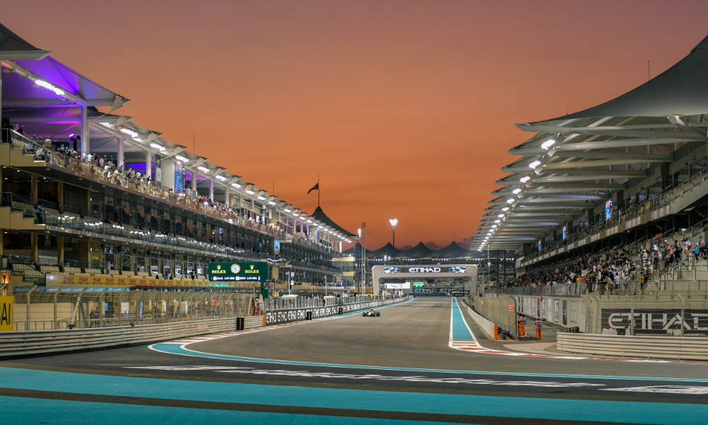 A view of a race track at sunset with grandstands filled with spectators on both sides, illuminated by bright lights, and mountains in the background.