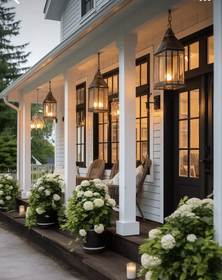 A front porch with white chairs, potted white flowers, and hanging lantern lights at dusk.