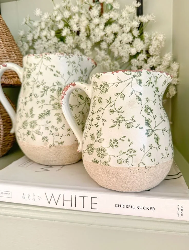 Two ceramic pitchers with floral patterns resting on a book titled "For the Love of White" by Chrissie Rucker, with a backdrop of white flowers.