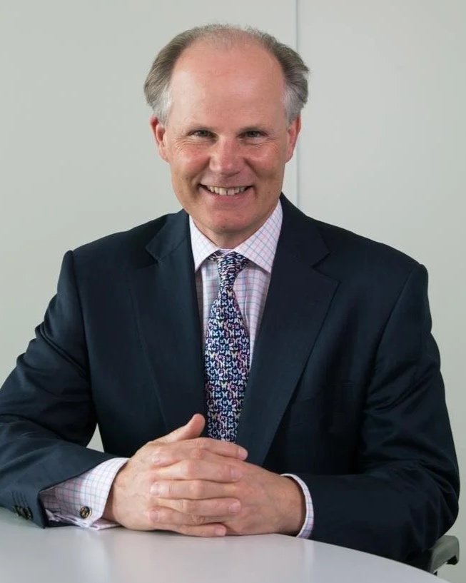 A smiling middle-aged man with gray hair, wearing a dark suit, checkered dress shirt, and a patterned tie, sitting at a desk with hands folded.