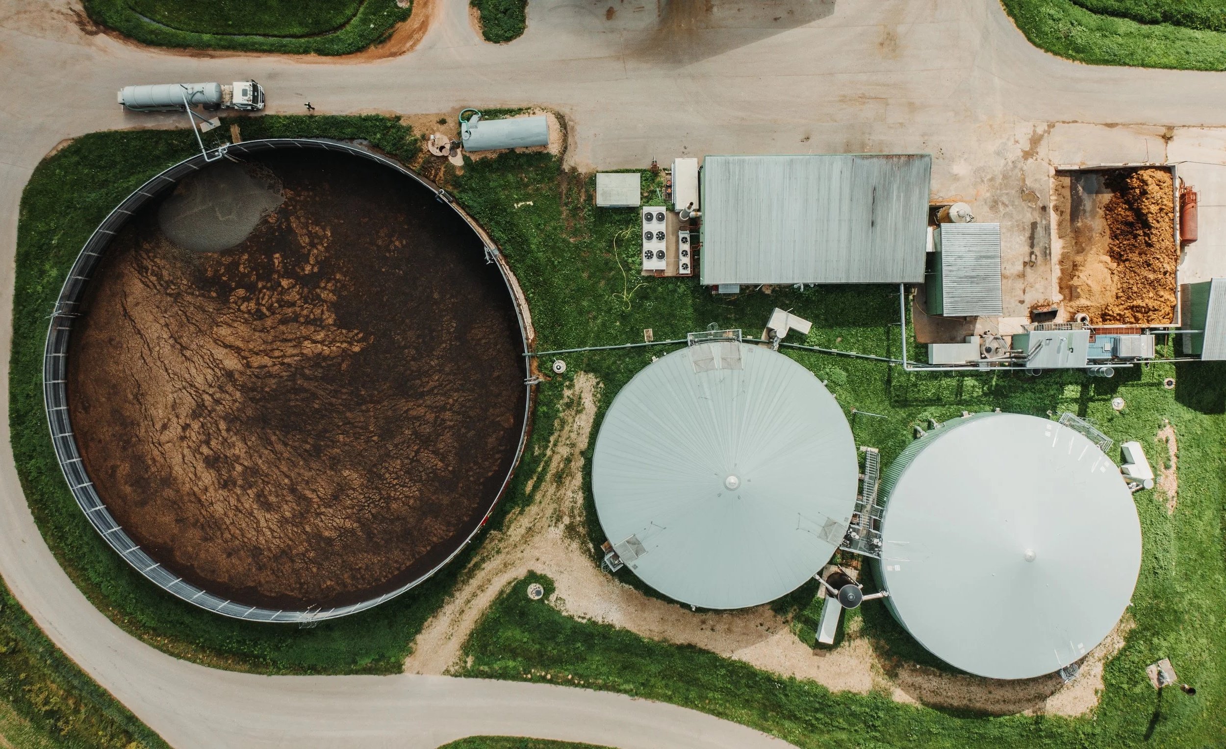An aerial view of wastewater treatment tanks and equipment, including large circular tanks with some containing sediment, surrounded by grassy areas, roads, and small structures.