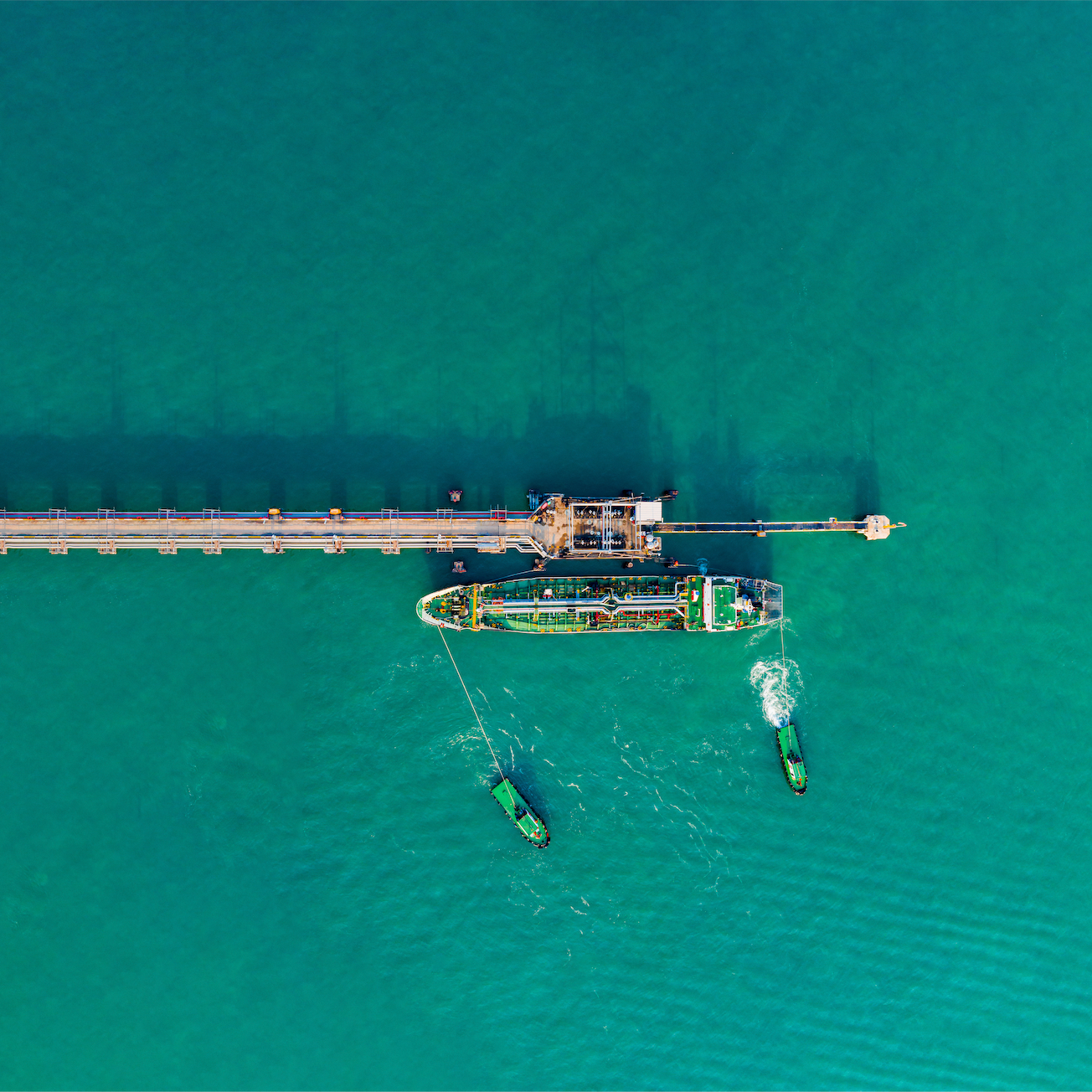 Aerial view of an oil or gas rig with two green tugboats nearby in turquoise water.