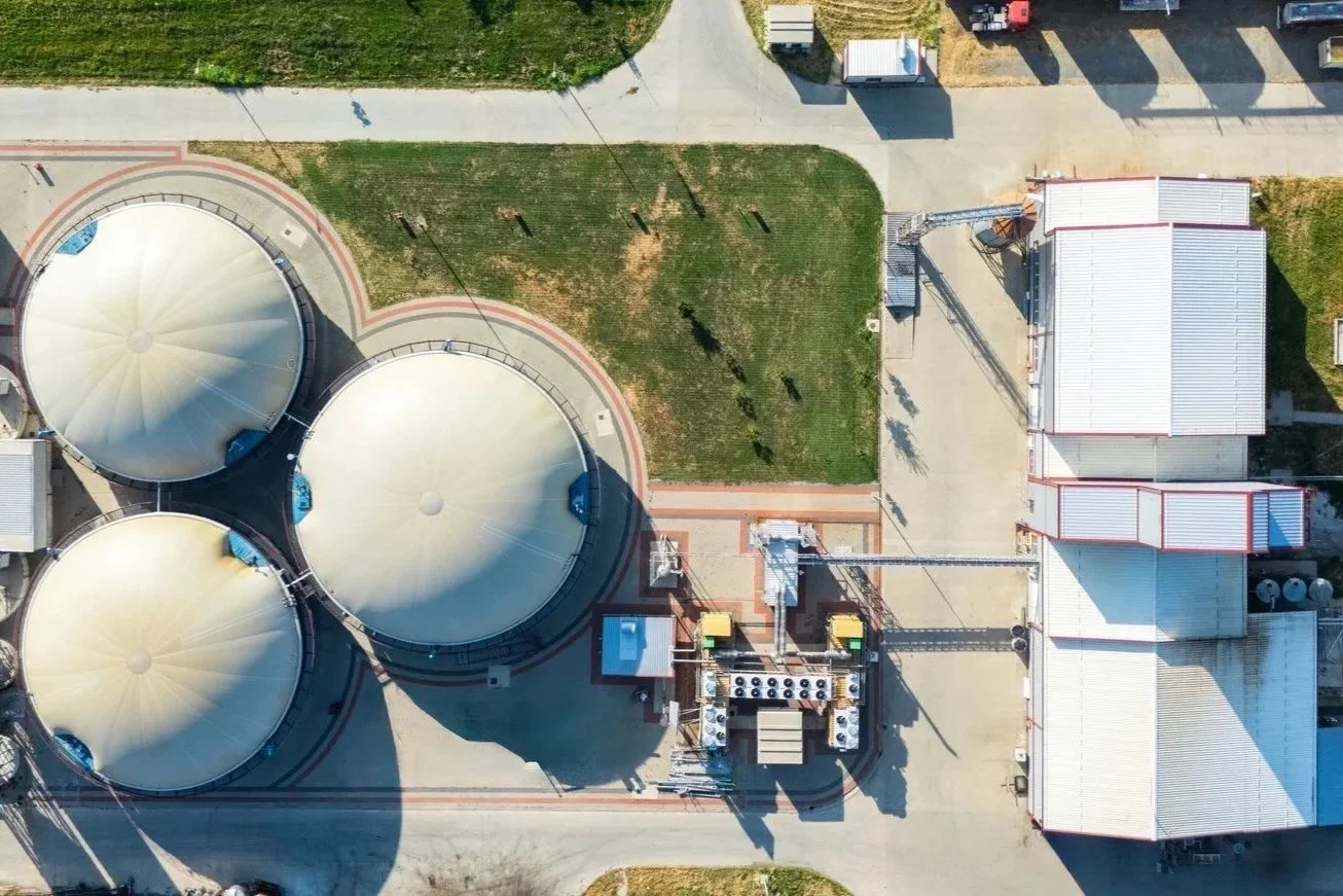 Aerial view of an industrial facility with three large storage tanks with cream-colored domed roofs, surrounded by paved pathways, grassy areas, and small buildings.