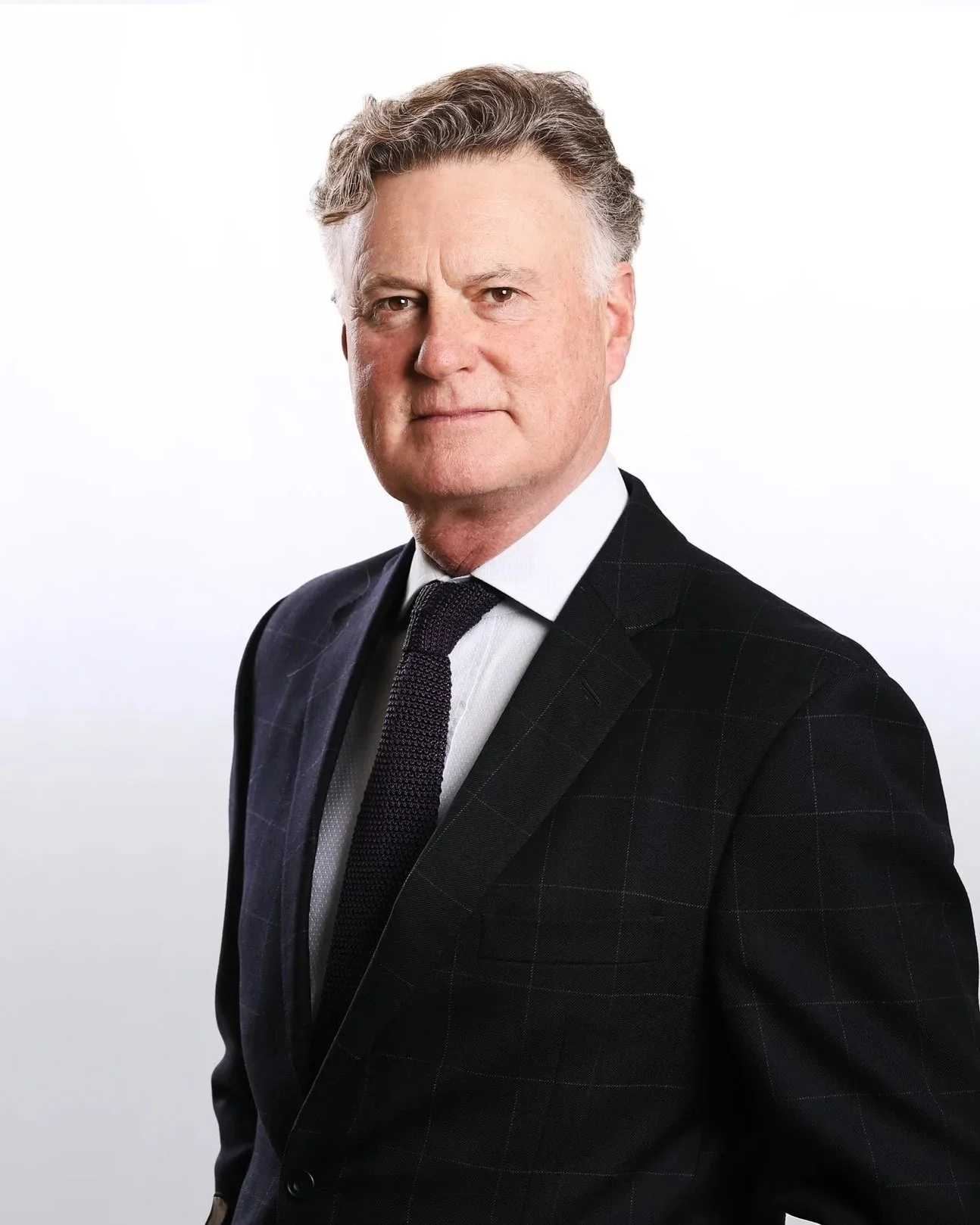 A middle-aged man with gray hair wearing a dark checkered suit, white shirt, and dark tie, looking confidently at the camera against a plain white background.