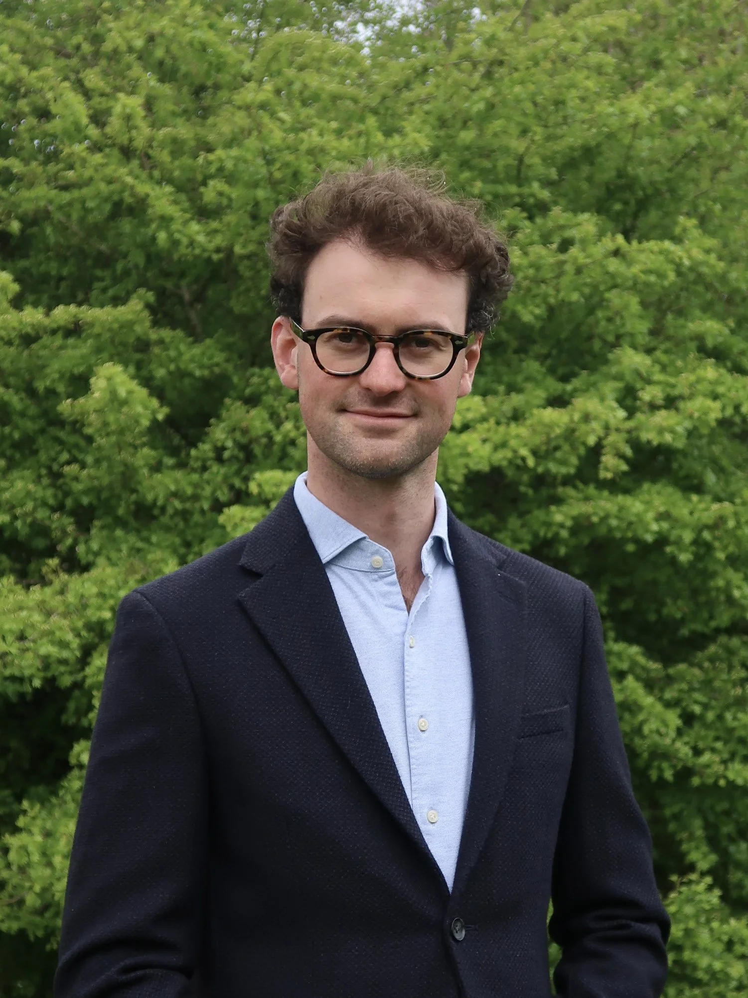 A man with brown curly hair and glasses wearing a navy blazer and light blue shirt standing outdoors in front of green leafy trees.