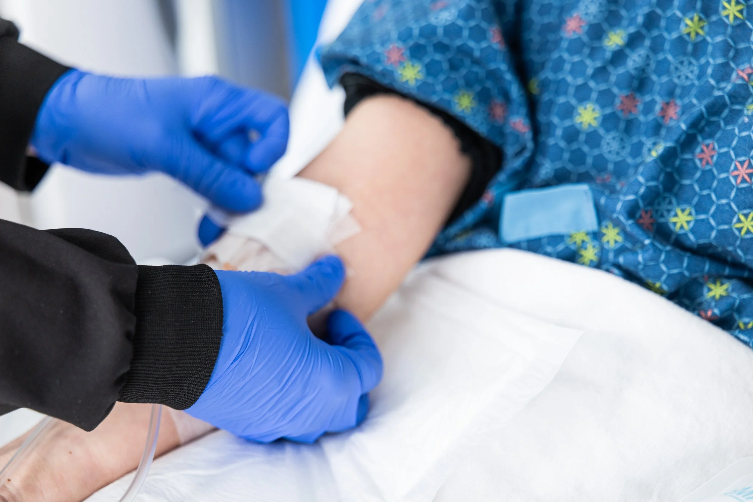 A healthcare worker wearing black long sleeves and blue gloves is drawing blood from a patient's arm. The patient is lying on a bed, wearing blue patterned clothing with colorful flowers, and has a white bandage on their arm.