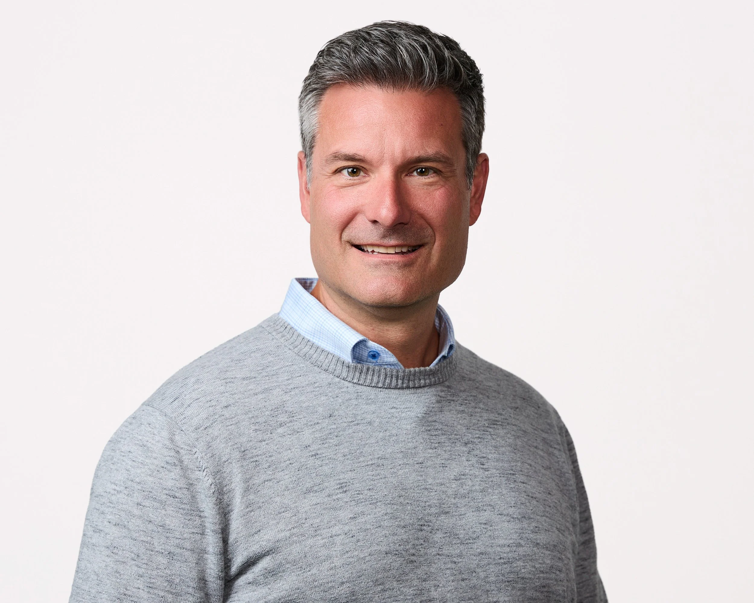 A middle-aged man with short, dark hair with some gray, wearing a light blue collared shirt and a gray sweater, smiling against a plain white background.