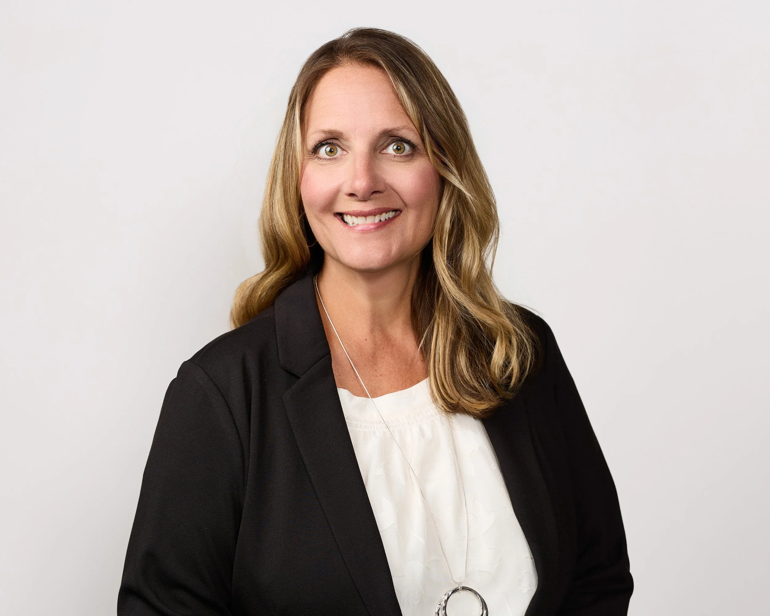 Portrait of a smiling woman with light brown hair, green eyes, wearing a black blazer over a white blouse, standing against a plain white background.