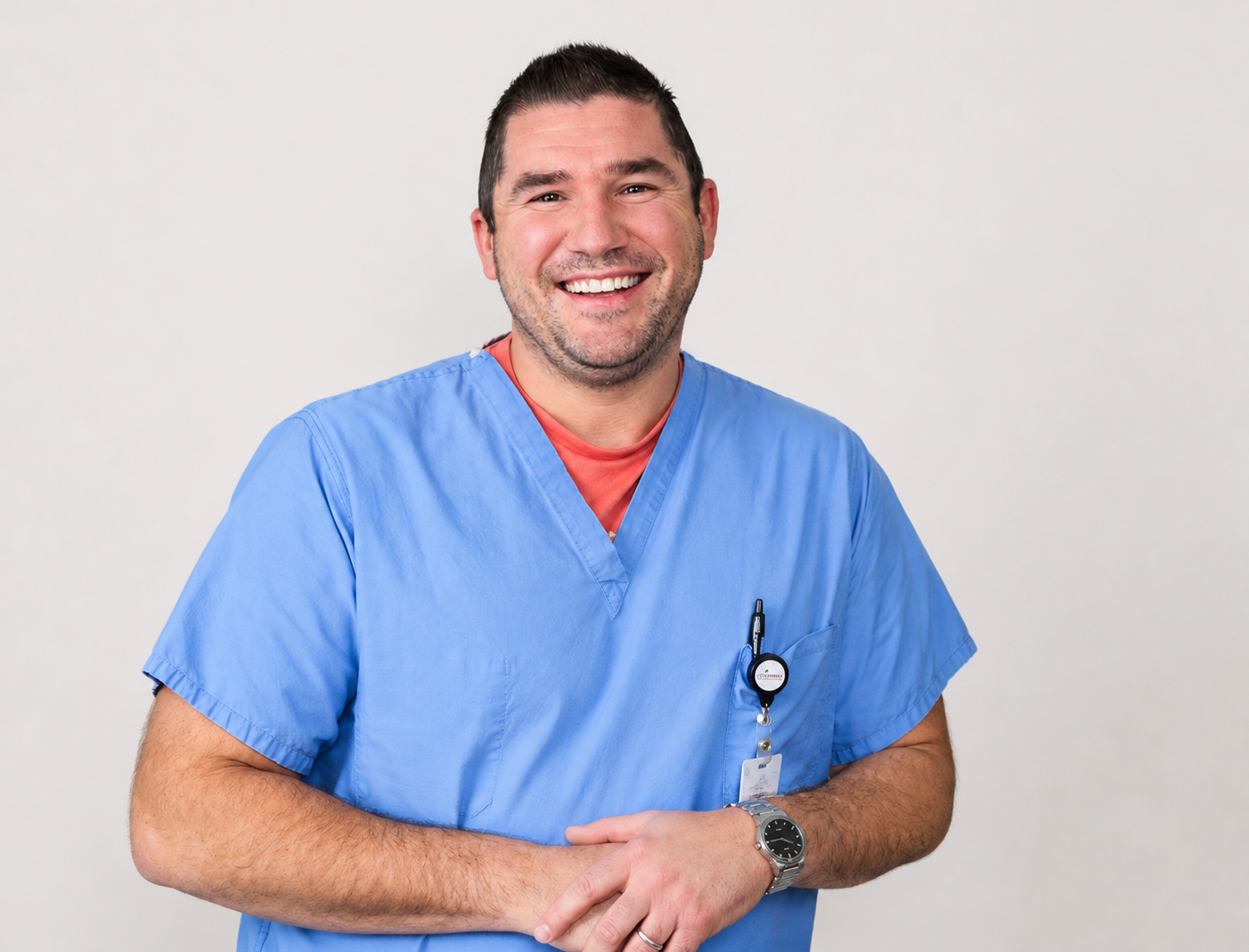 A smiling male healthcare professional in blue scrubs standing against a plain background.