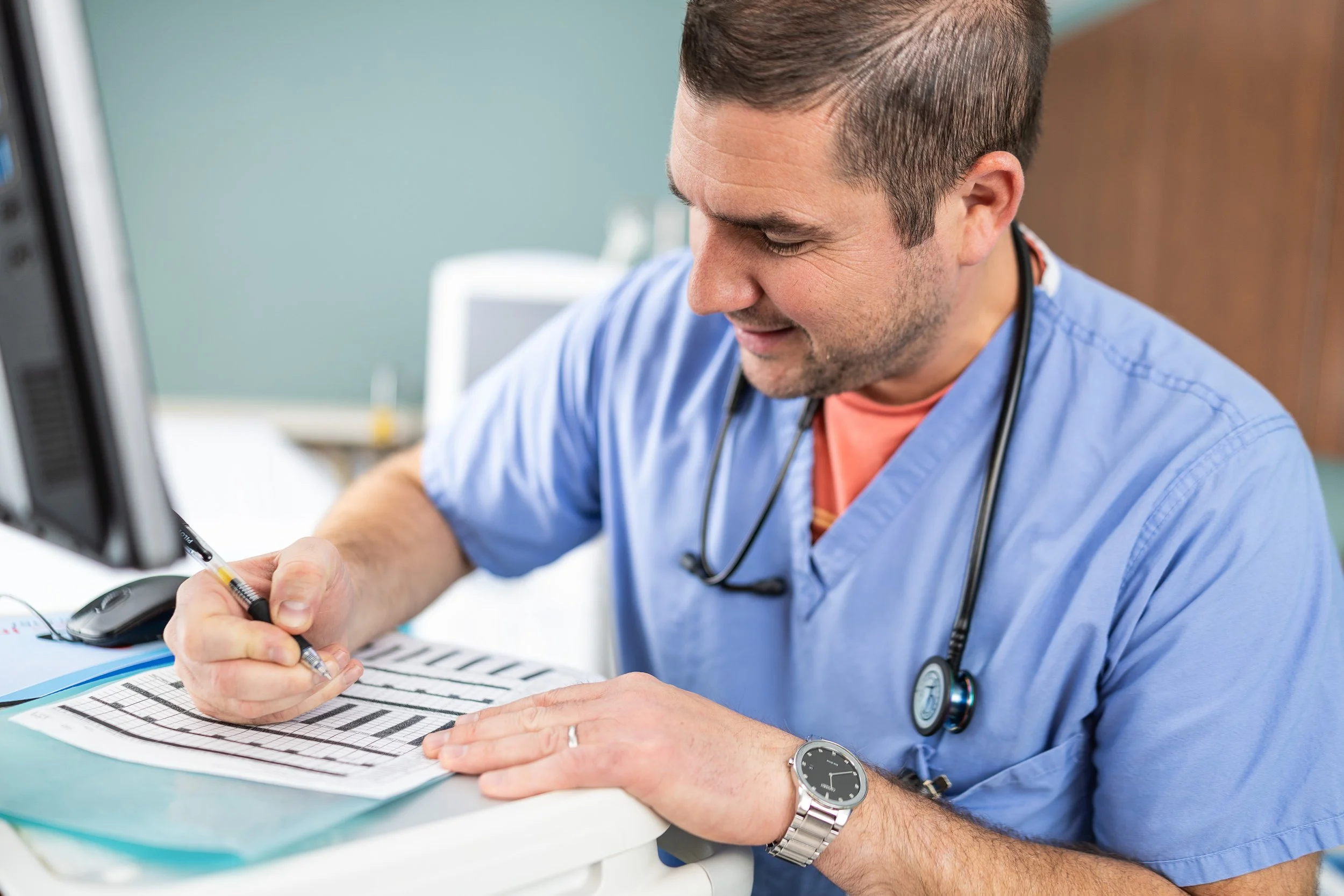 A male healthcare professional in scrubs and a stethoscope around his neck writing on medical paperwork with a pen, sitting at a desk in a medical setting.