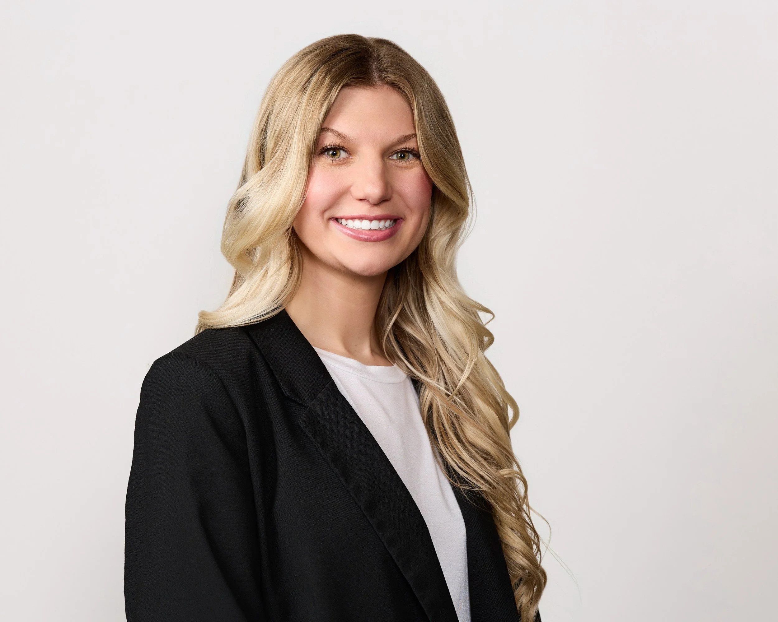 A woman with long blonde wavy hair, wearing a black blazer and a white T-shirt, smiling against a plain white background.