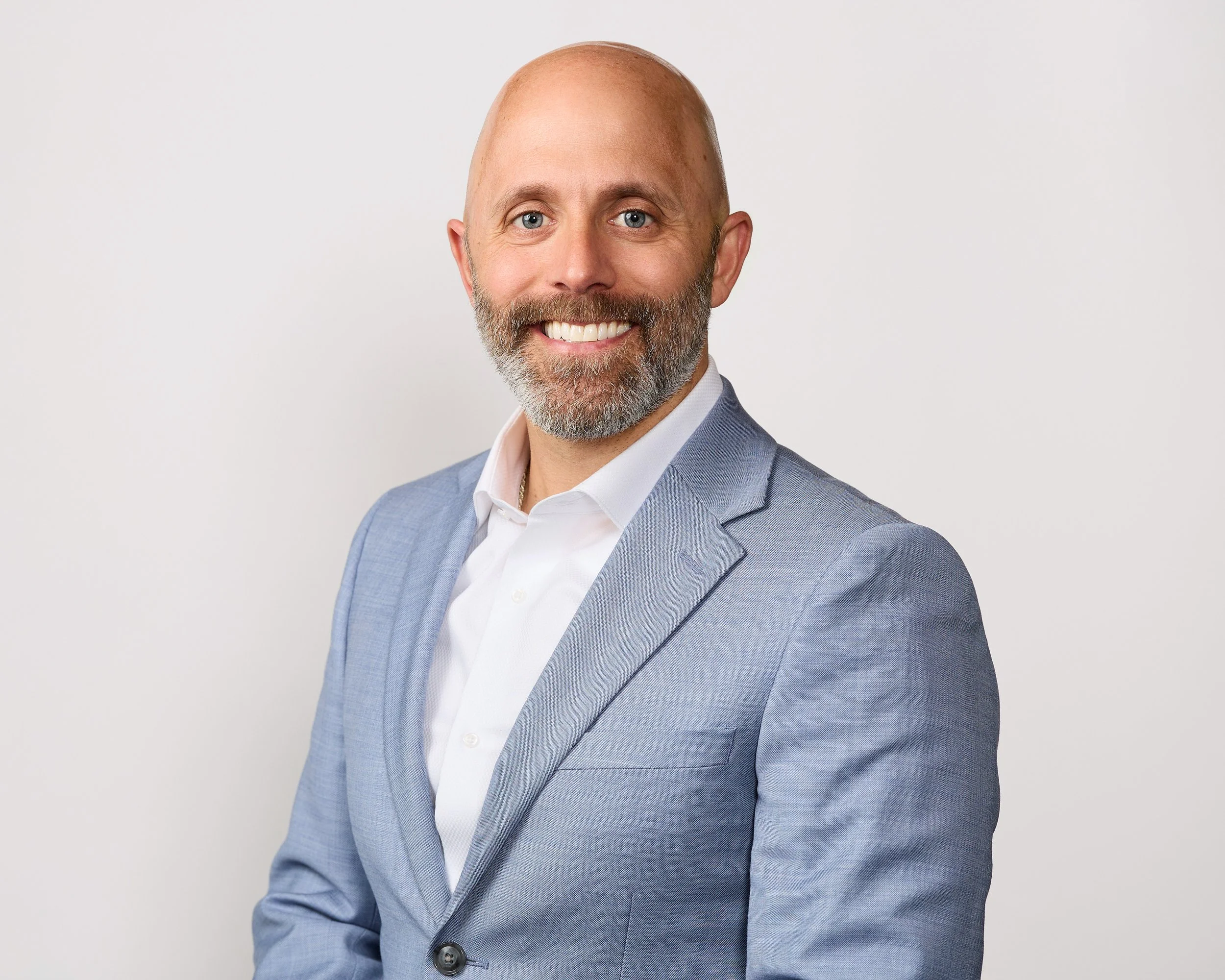 A man with a bald head and a beard, smiling, wearing a light blue suit and white shirt, posing against a plain white background.
