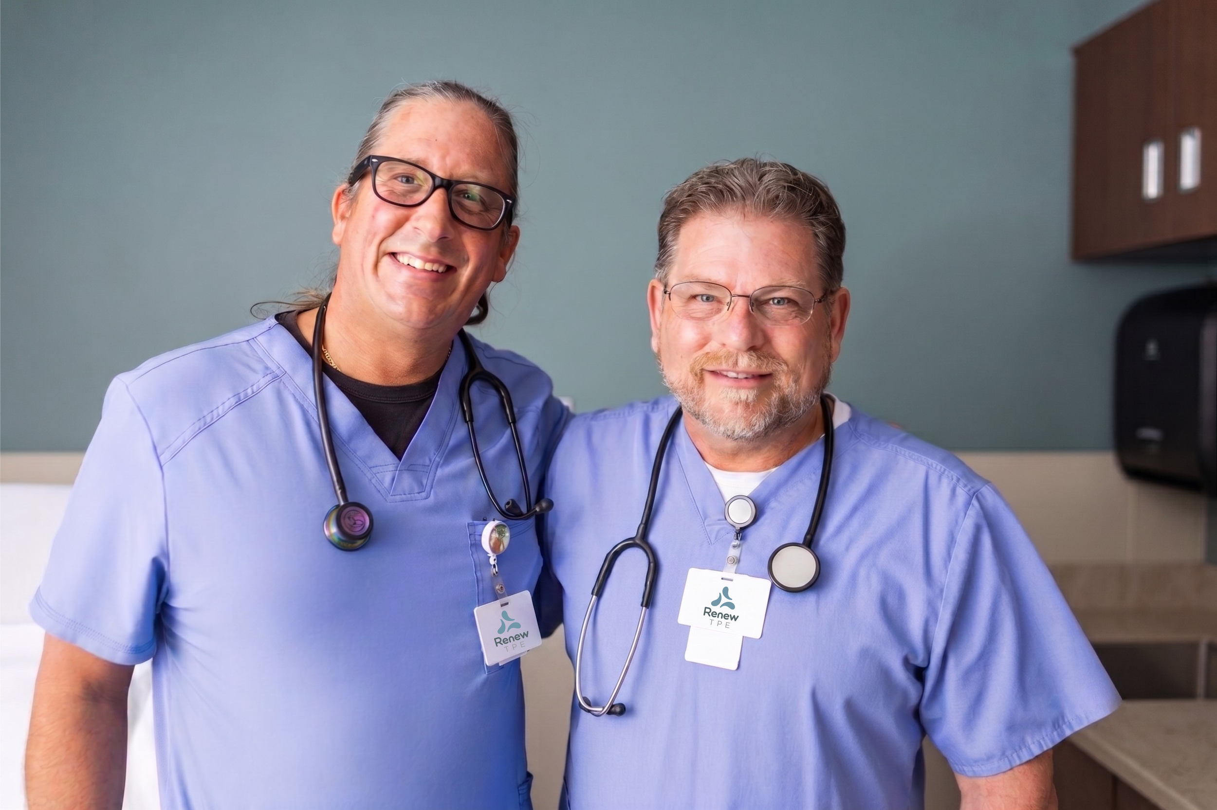 Two male healthcare workers in blue scrubs smiling and posing for a photo in a medical setting, with attached stethoscopes and identification badges.