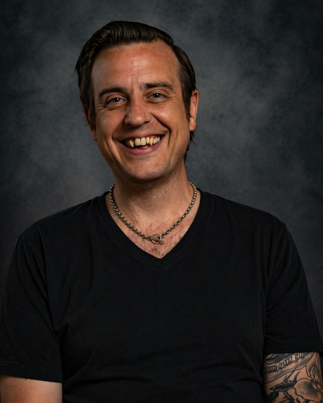 A smiling man with dark hair, a chain necklace, wearing a black V-neck shirt, and has tattoos on his arm, against a dark textured background.