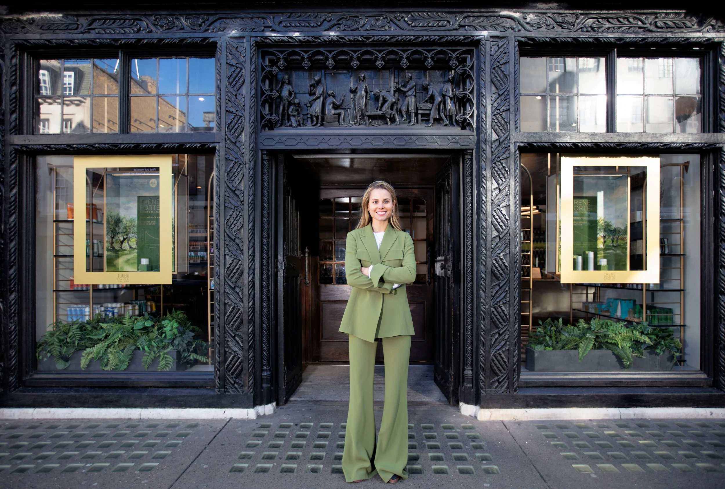 A woman in a light green suit standing in front of a dark, ornate building entrance with glass display windows on either side, greenery at her feet, and a carved bas-relief sculpture above the doorway.