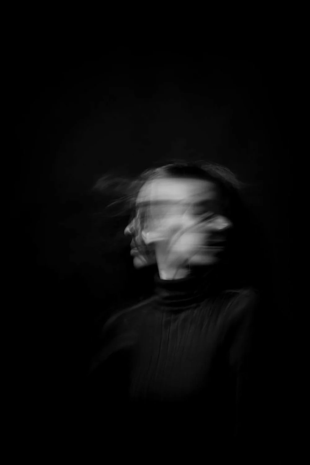 A blurred black-and-white photo of a woman with her hair being blown in a dark setting.