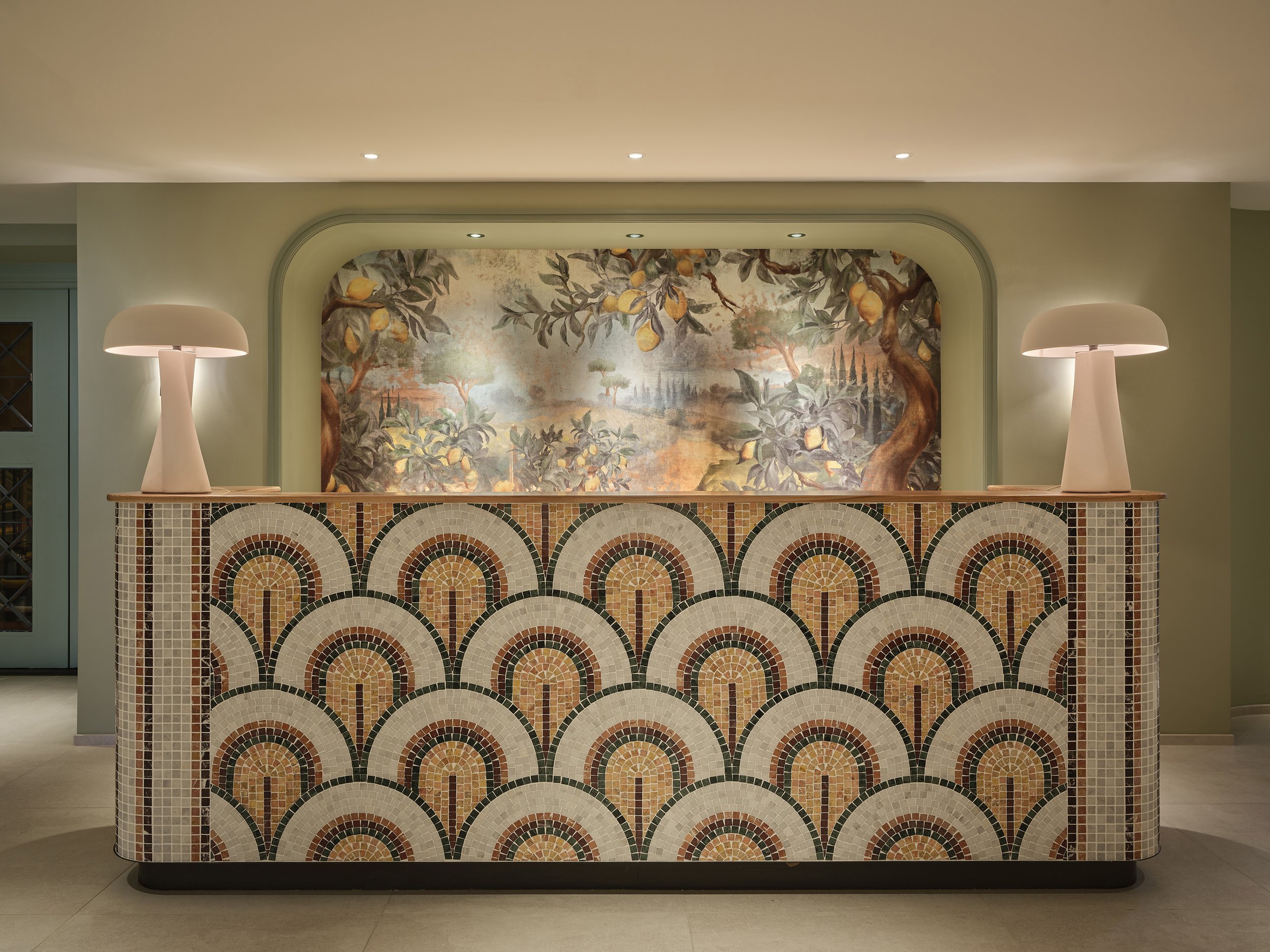 A mosaic front desk with a rounded arch design, topped with two white lamps, in front of a painted landscape mural featuring trees and foliage.