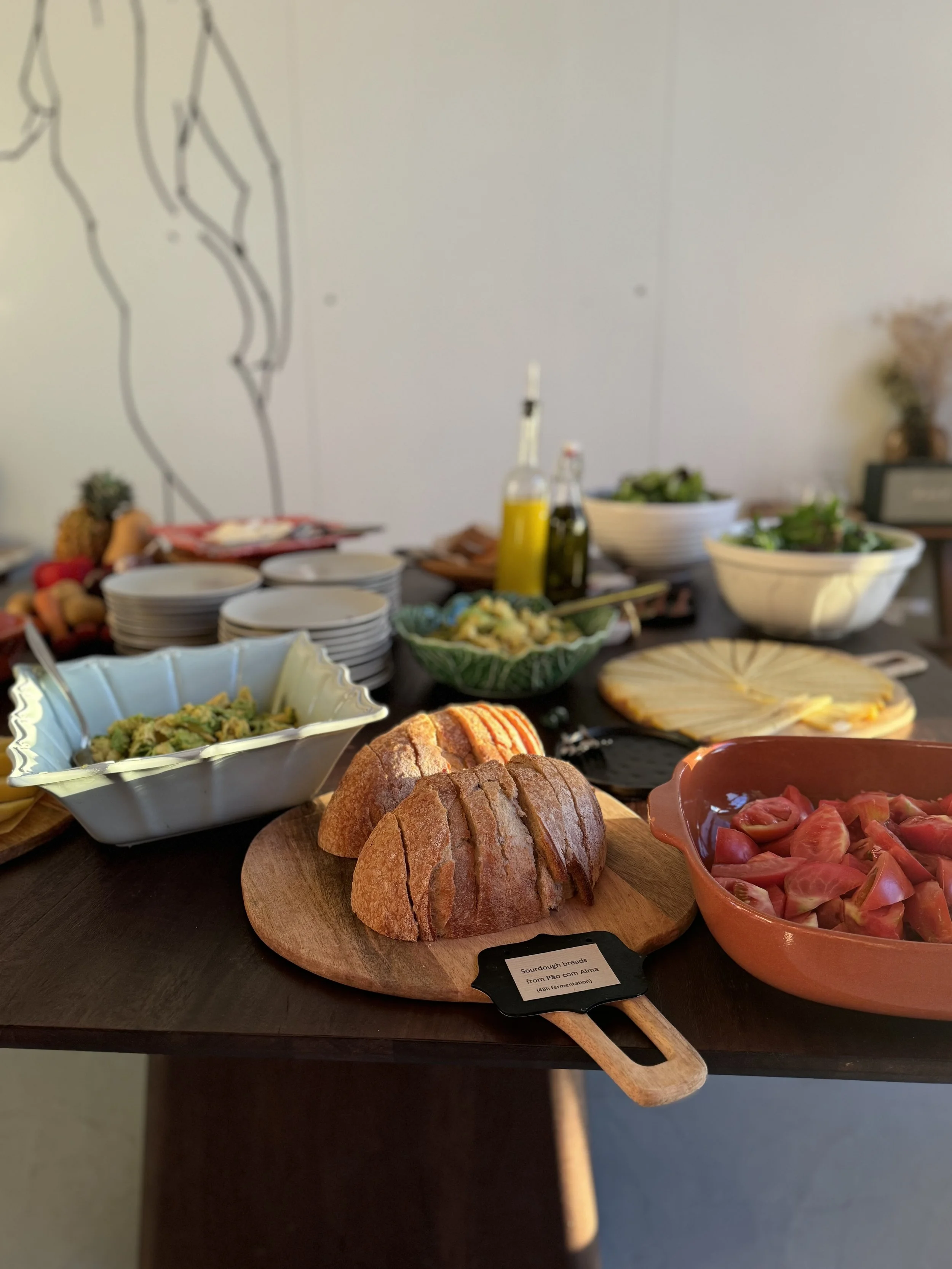 Sourdough bread slices on a wooden cutting board with a label that reads "Sourdough breads from Pao con Alma". Surrounding dishes include chopped tomatoes in an orange bowl, a bowl of salad, bowls of greens, and bottles of oil and vinegar. In the bac