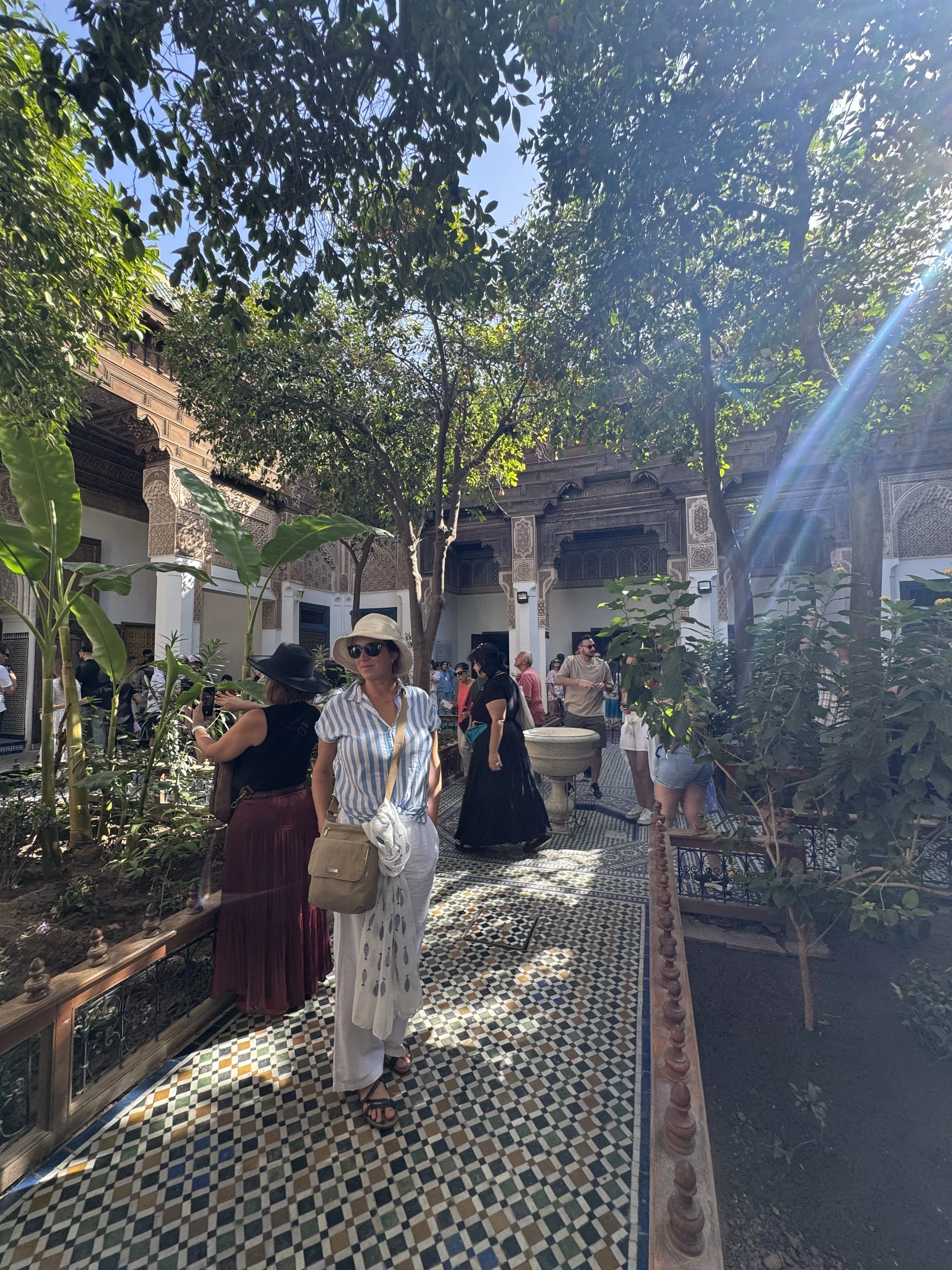 People walking and standing in a courtyard with patterned tile floor, surrounded by lush greenery and ornate building architecture, sunlight streaming through trees.