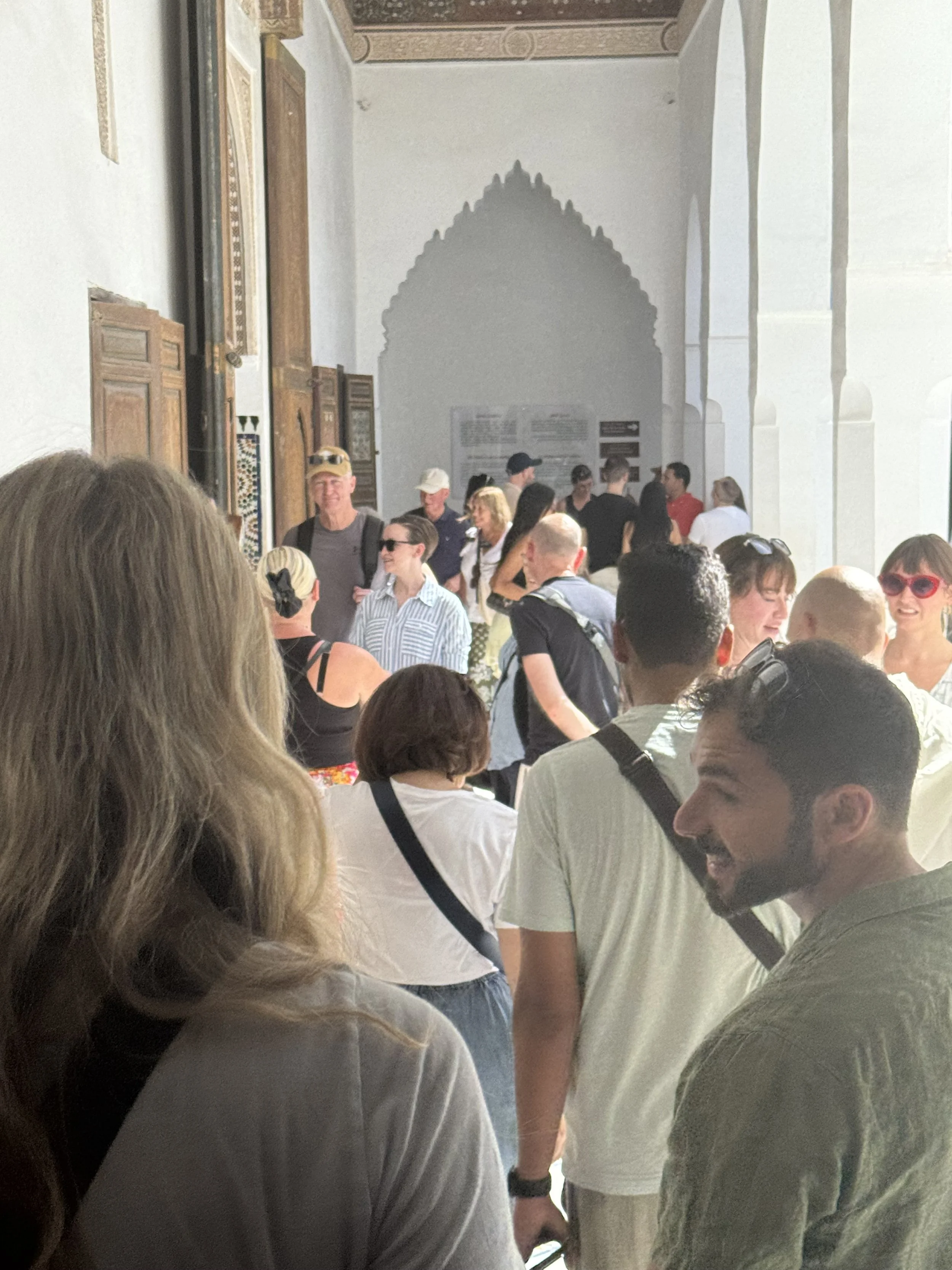 A crowded indoor space with many people standing and talking. The background has arches and decorative elements on the wall.