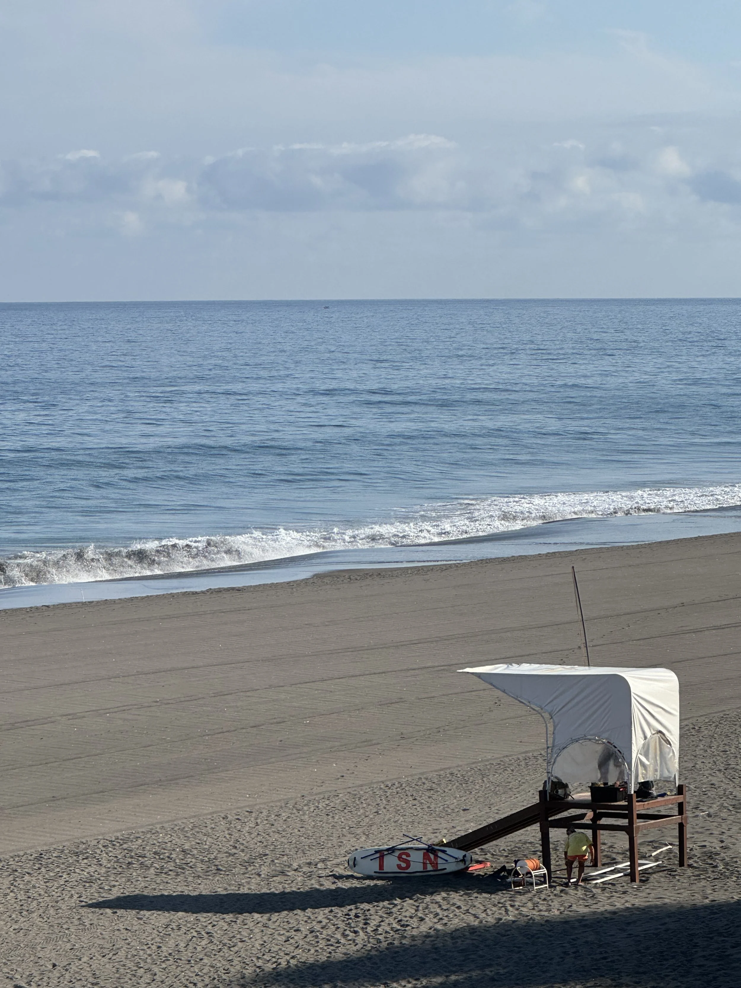 A sandy beach with a white sailboat in the foreground, a lifeguard stand with a white canopy, and a woman in a yellow top near the stand. The ocean has gentle waves, and the sky is partly cloudy.