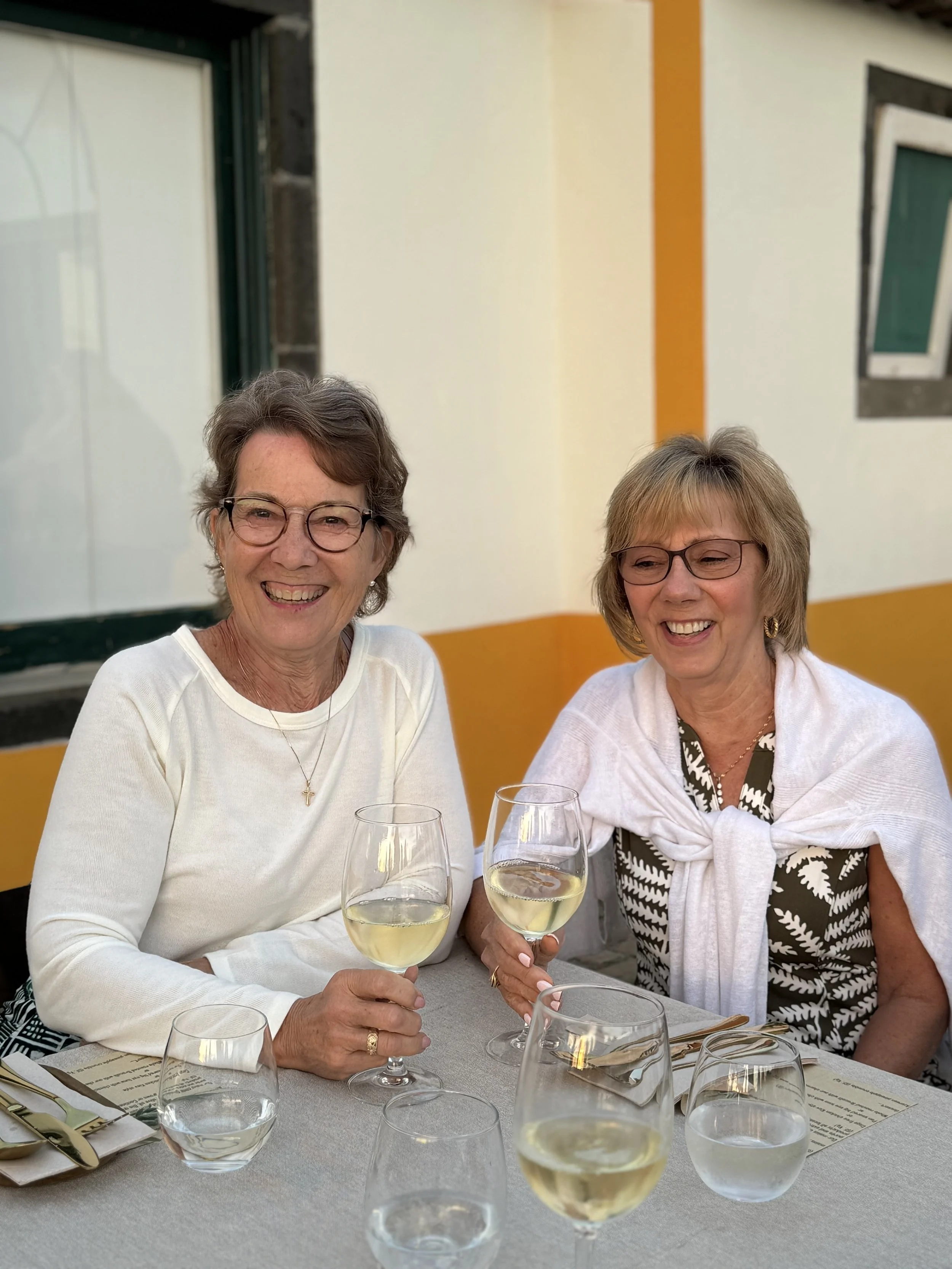 Two women smiling and holding glasses of white wine at a table set with glasses, silverware, and menus, outdoors at a restaurant with yellow and white walls.