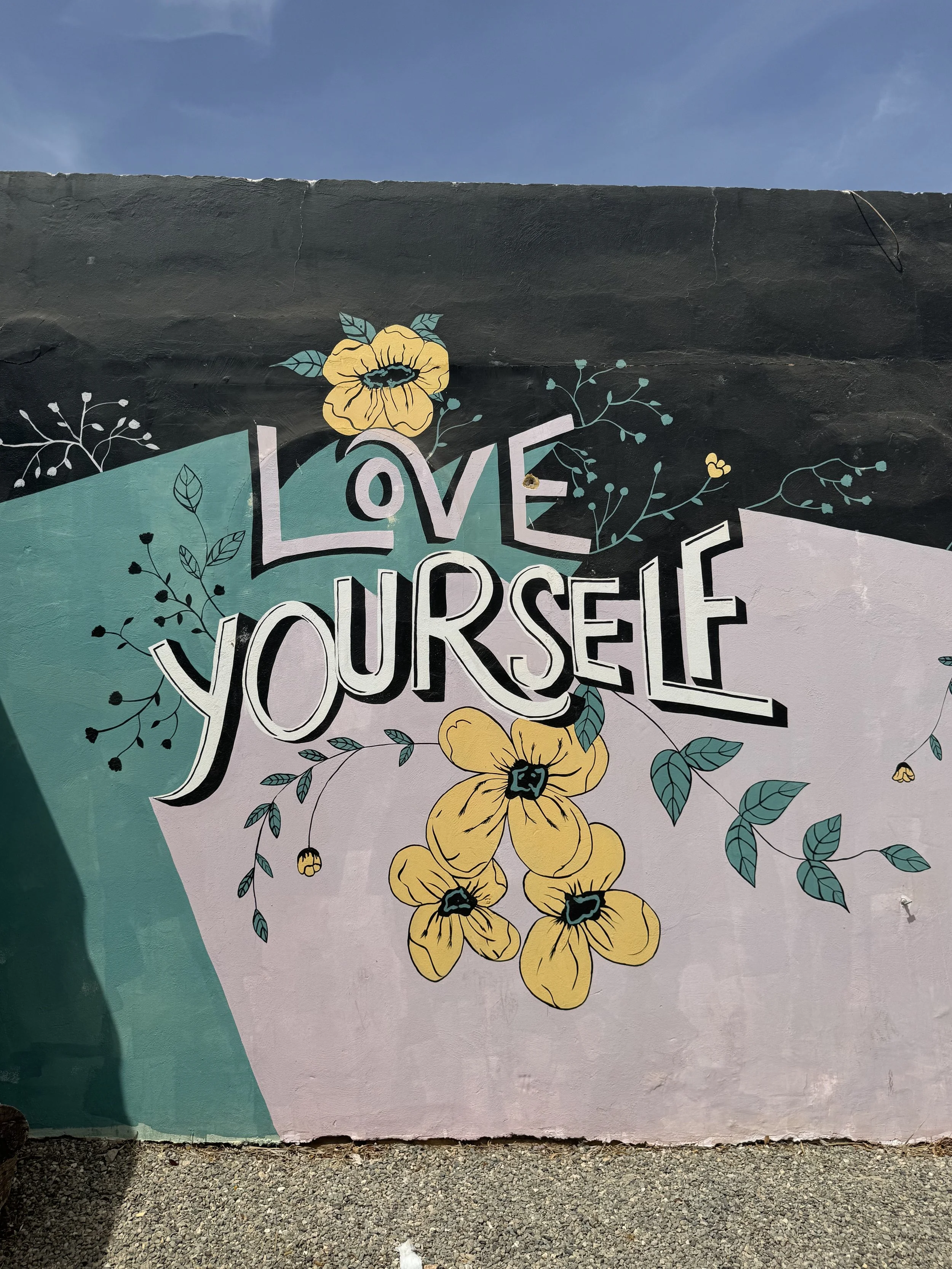 A mural on a wall with the words 'Love Yourself' surrounded by yellow flowers and green leaves, with a blue sky above.