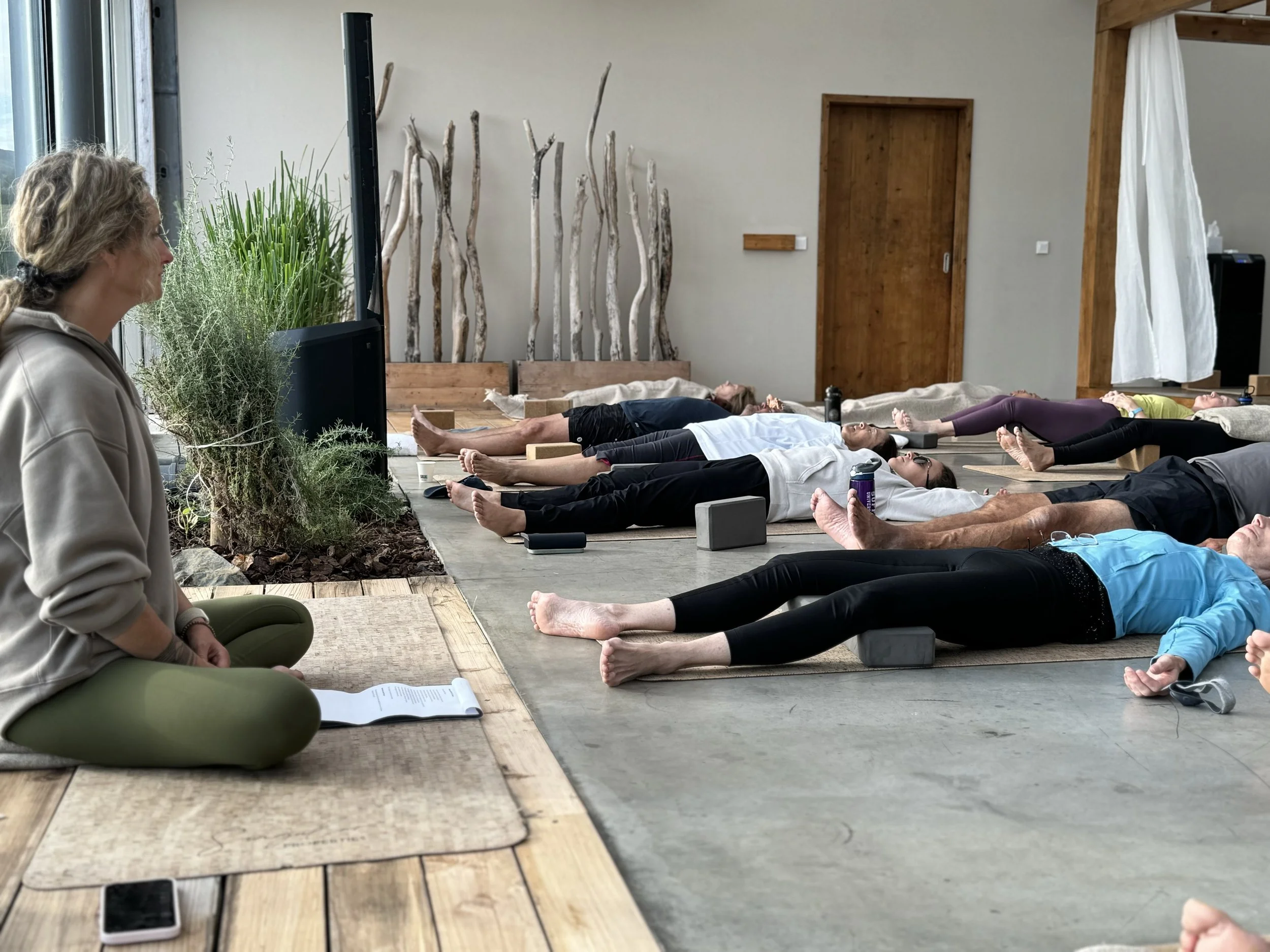 A group of people practicing yoga indoors, lying on mats with blocks, with a woman in the foreground observing. The room has large windows, plants, and wooden accents.