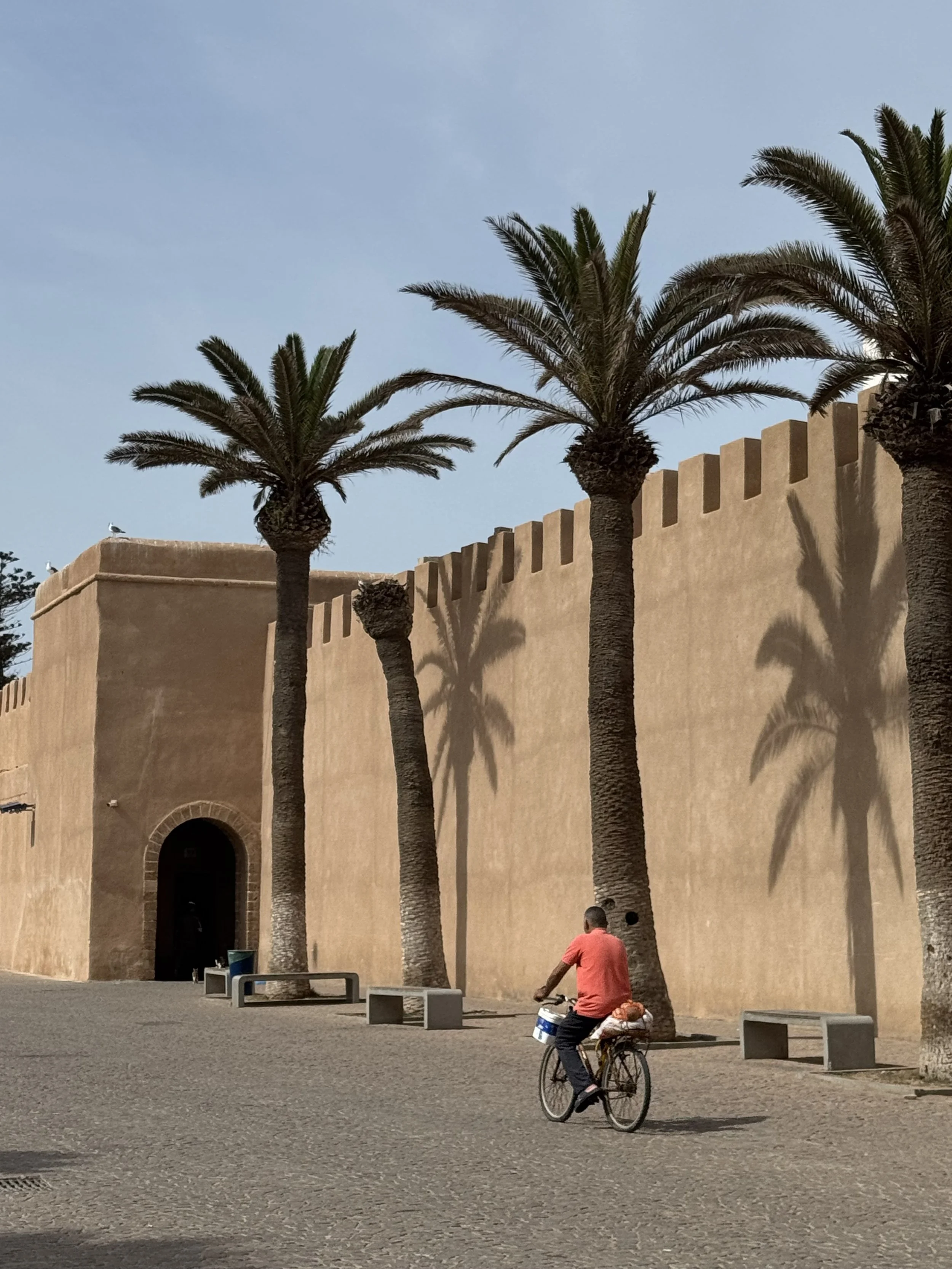 A man wearing a red shirt riding a bicycle in front of a beige building with palm trees and a clear blue sky.