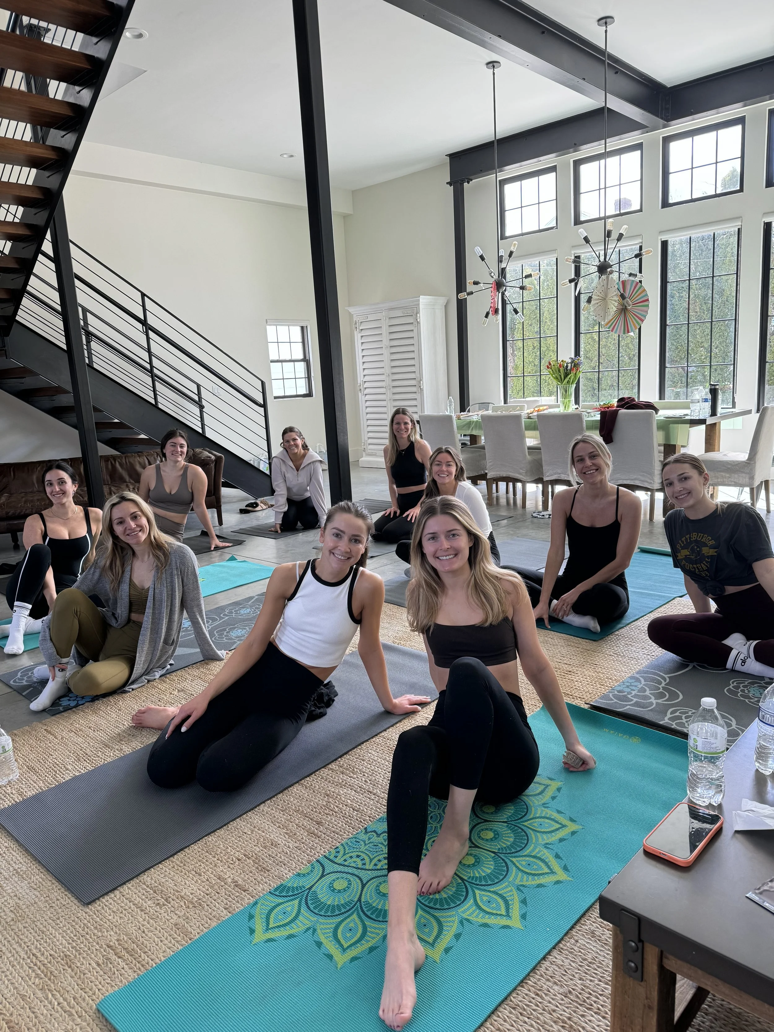 A group of women participating in a yoga or fitness class in a bright, spacious room with large windows, some sitting on yoga mats, smiling at the camera.