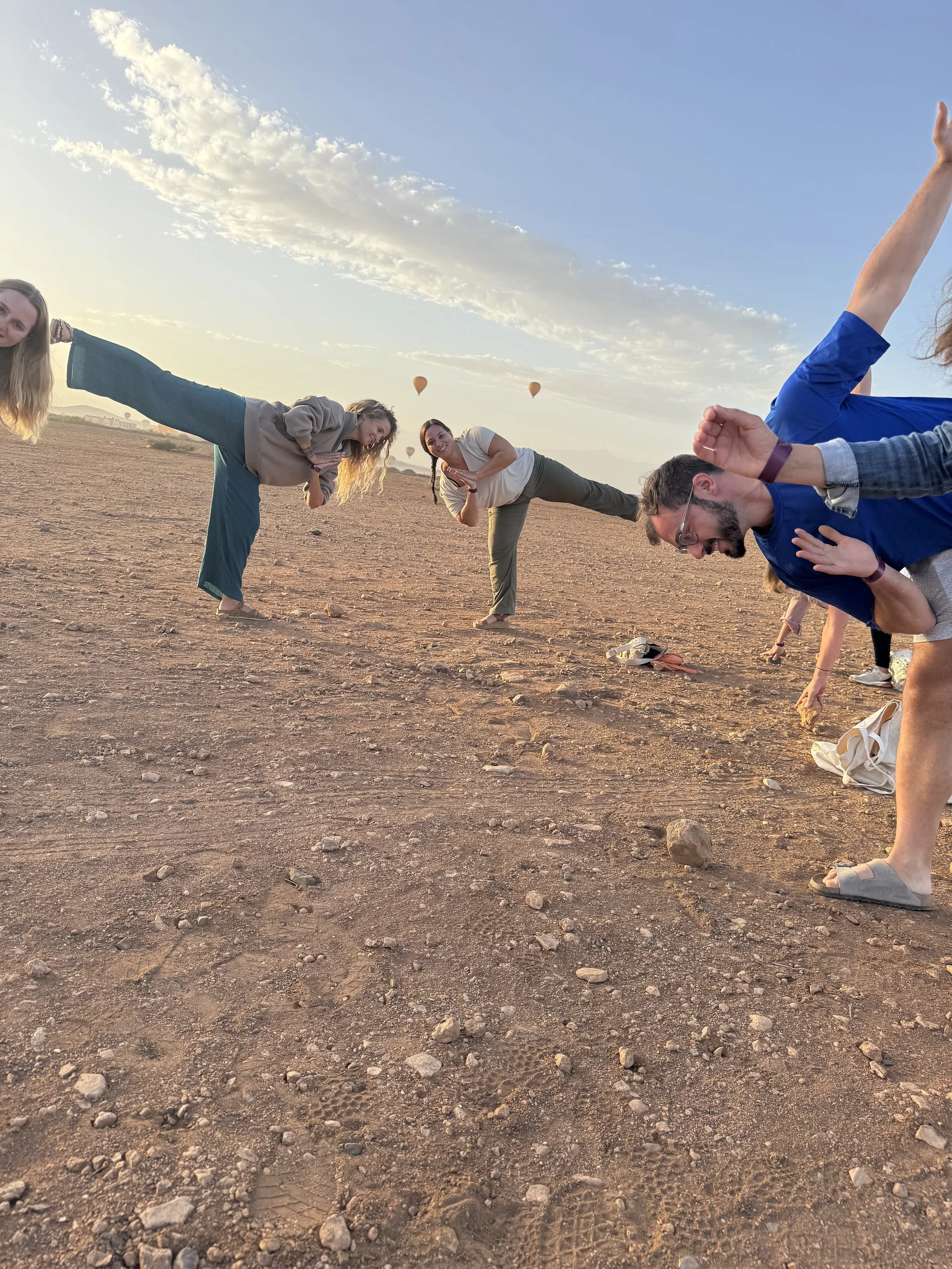 Group of people doing yoga poses on desert ground during sunset, with hot air balloons in the sky.