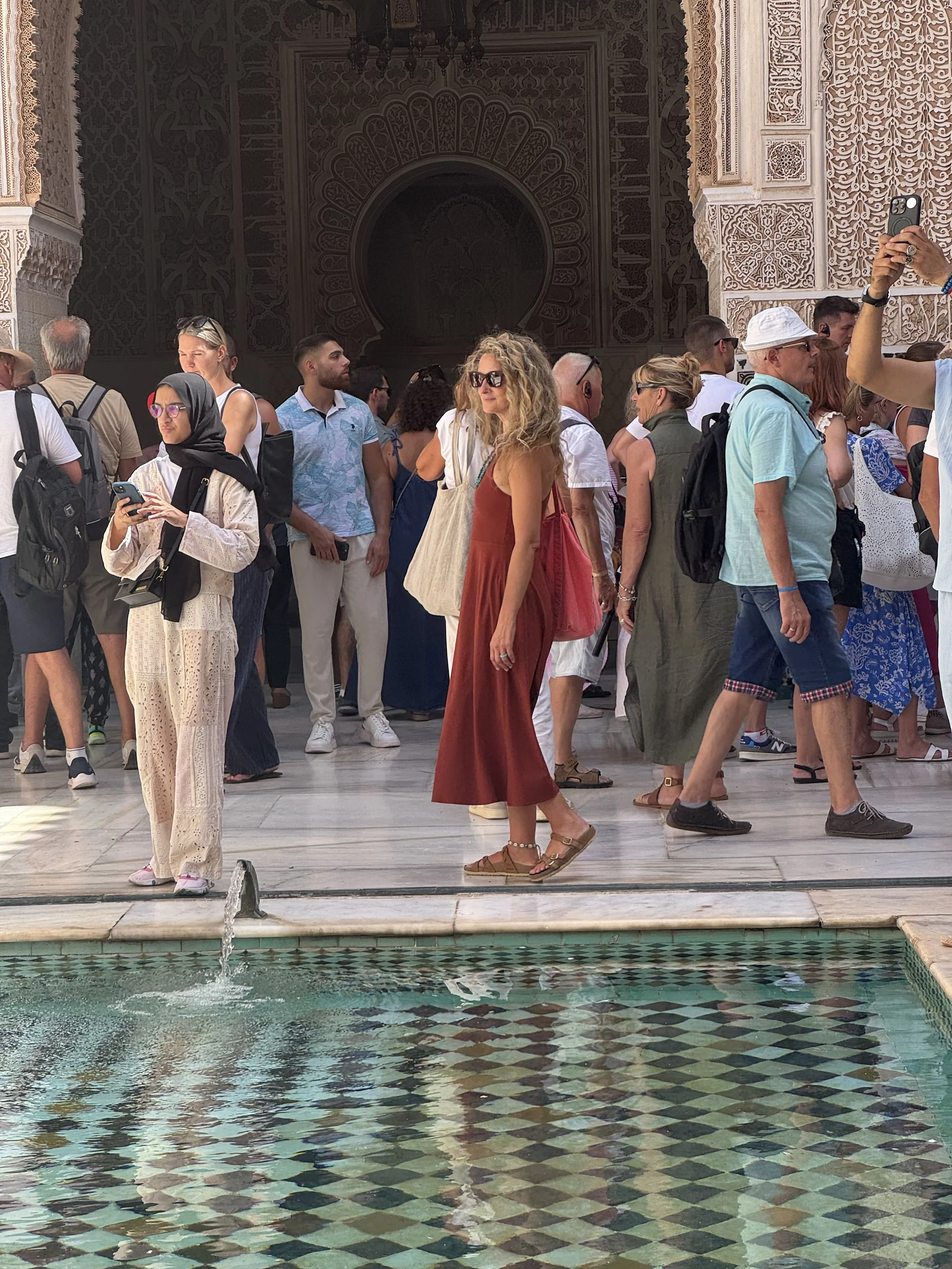 A group of tourists standing on the edge of a fountain inside a Moroccan palace, with intricate arabesque designs on the walls behind them.