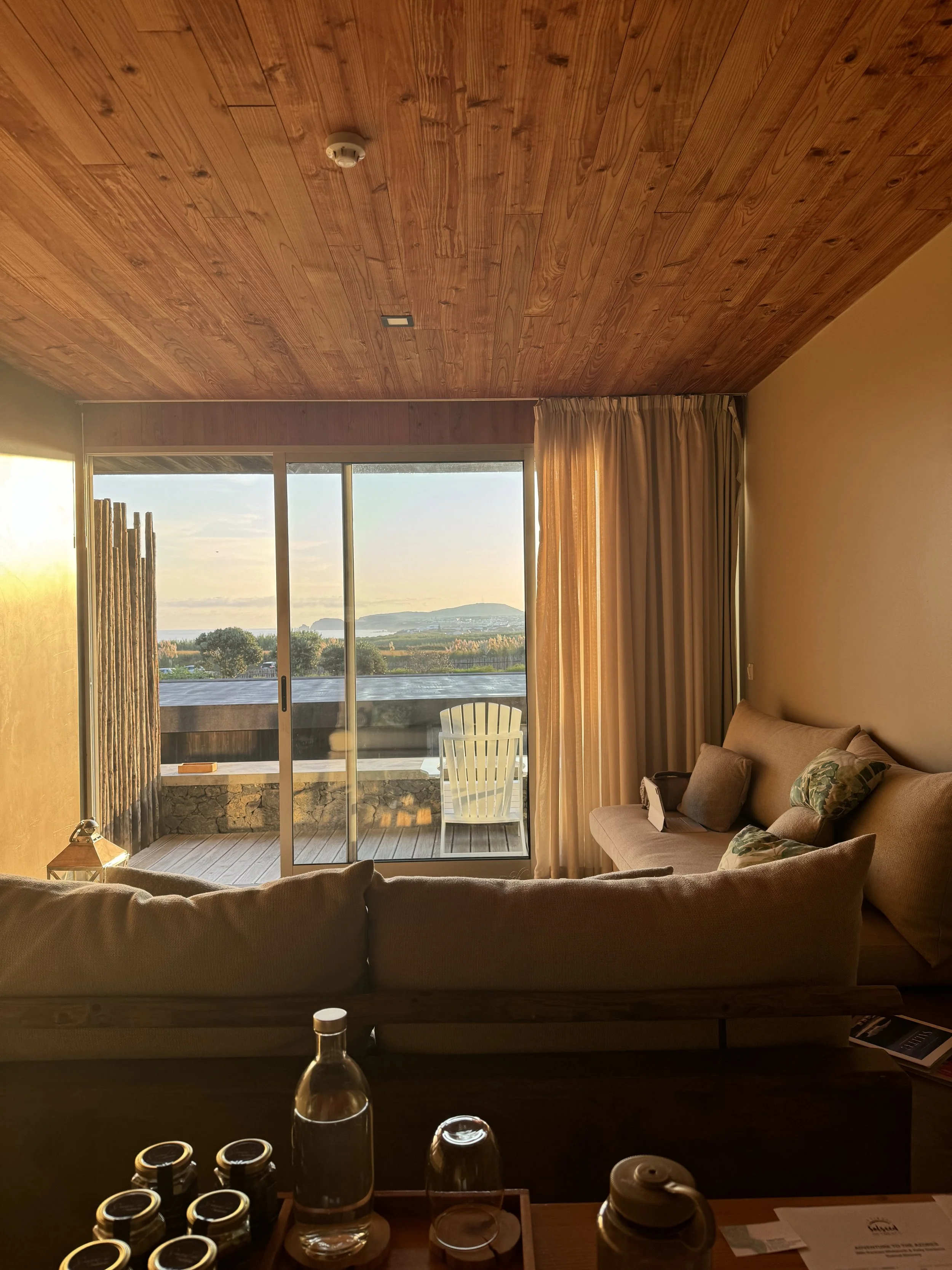 Living room with a large window showing a scenic view of mountains and water, beige sofa with pillows, wooden ceiling, curtains, and outdoor balcony with a chair.