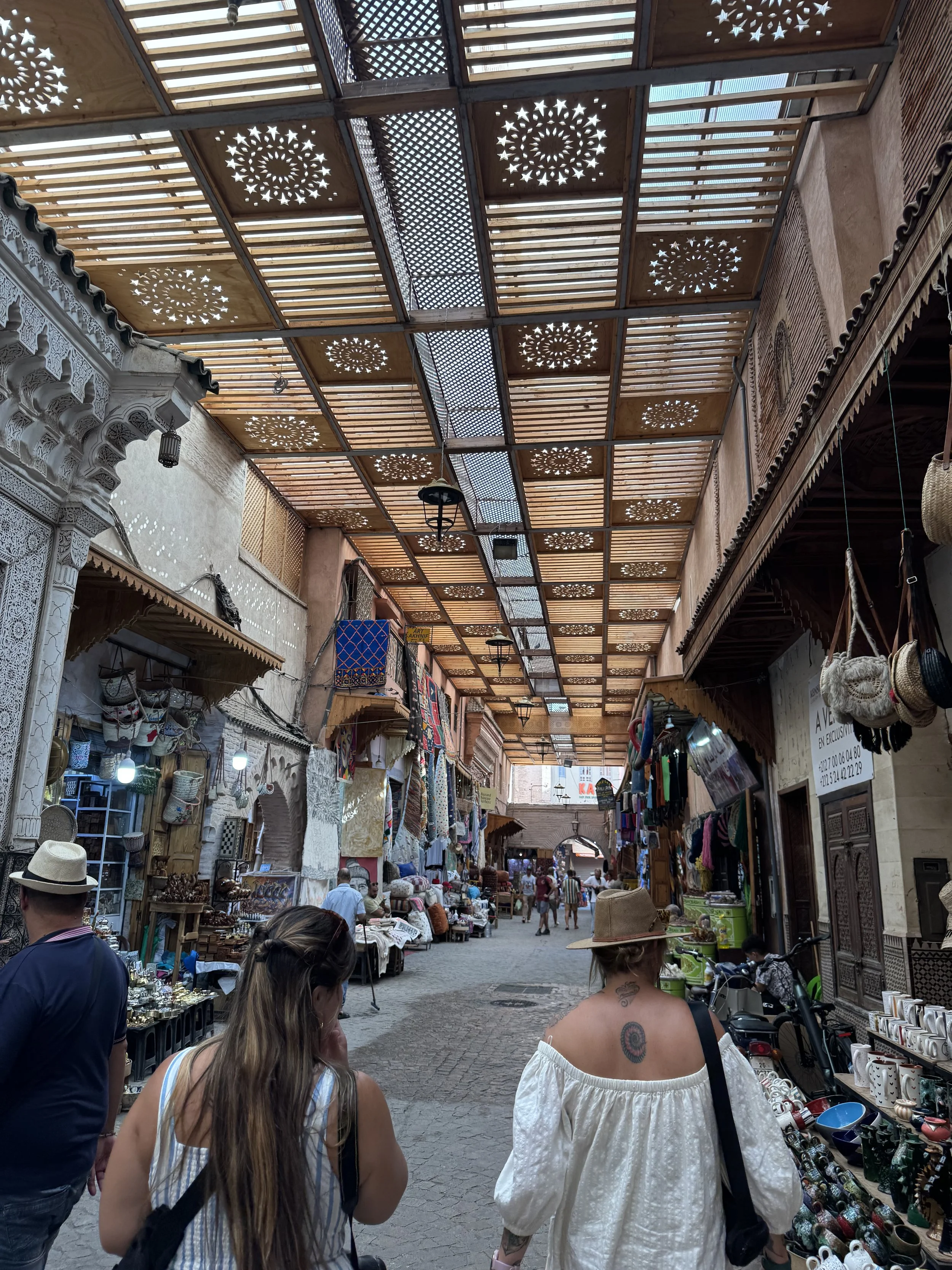Outdoor marketplace with stalls selling textiles, jewelry, and ceramics. Covered with a decorative wooden roof featuring star and circle cutouts. Shoppers browsing and walking along cobblestone street.