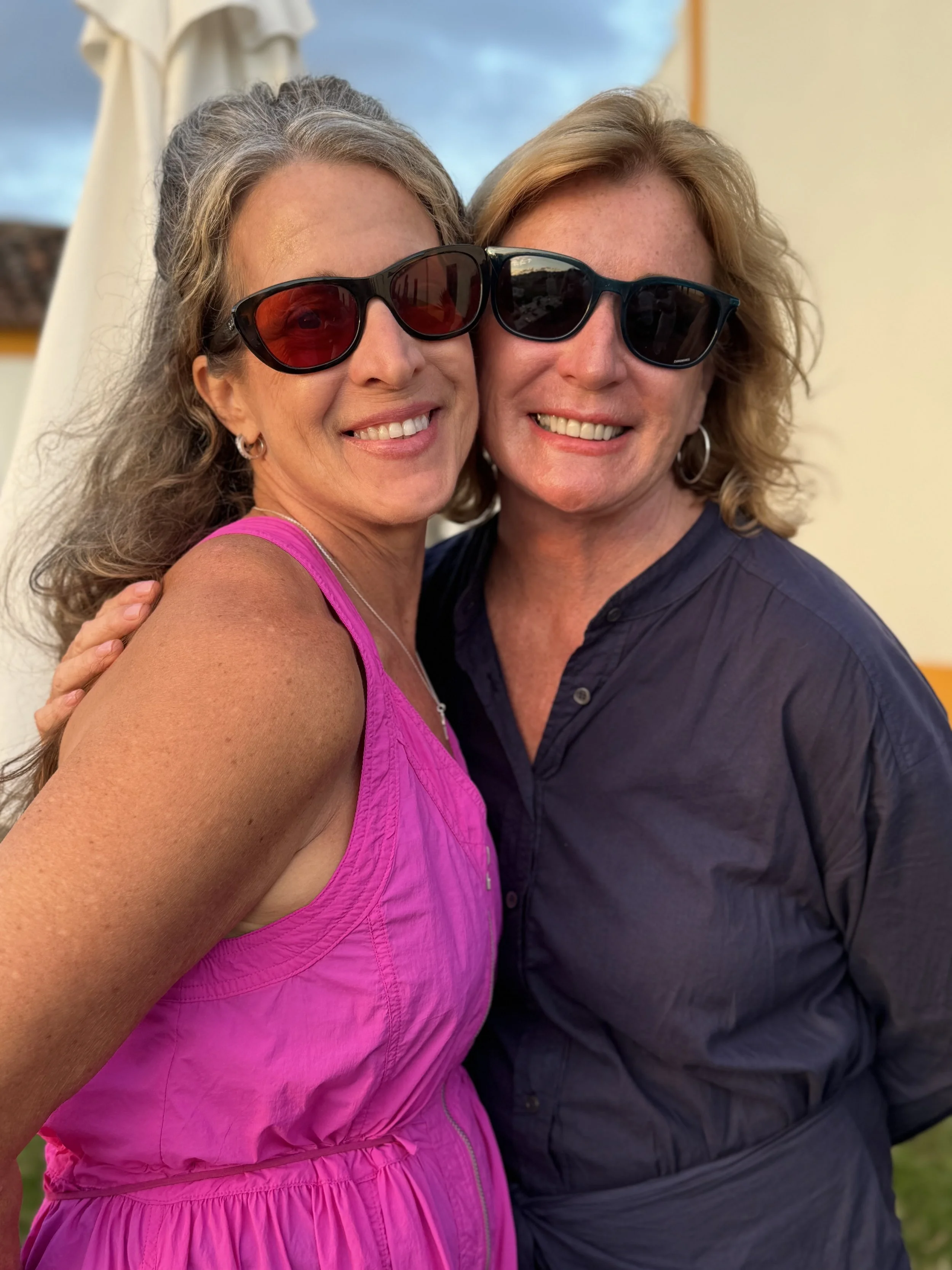 Two women wearing sunglasses, smiling and hugging, outdoors on a bright day.