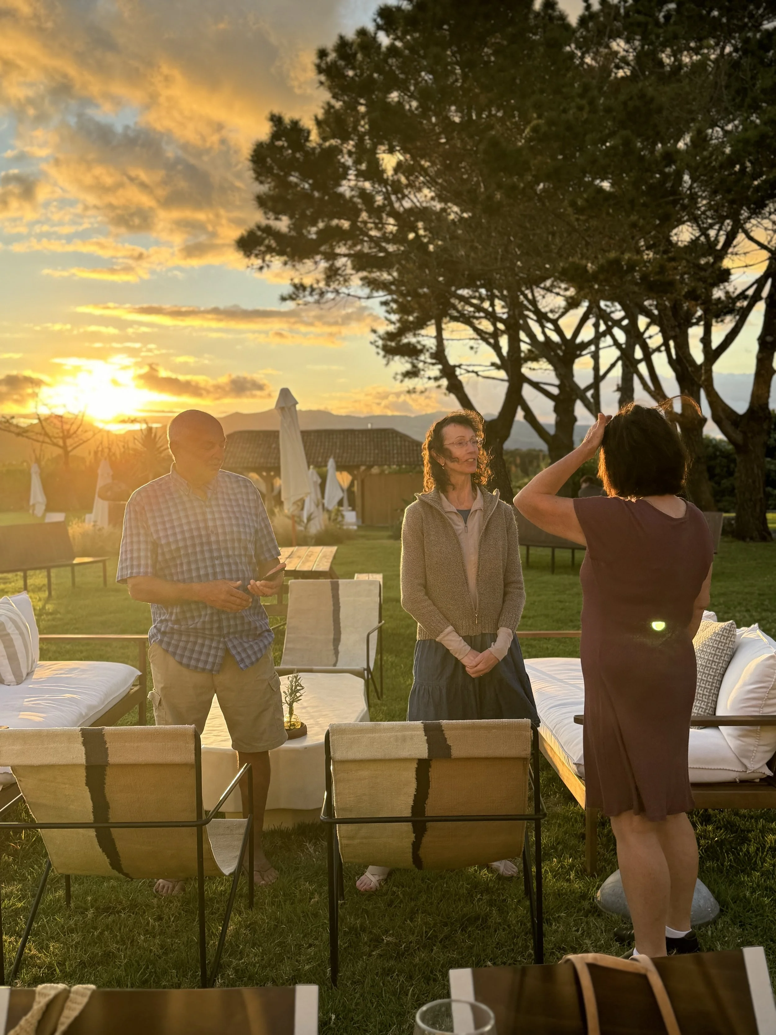 Three people engaged in conversation during sunset in an outdoor setting with tables, chairs, trees, and a house in the background.