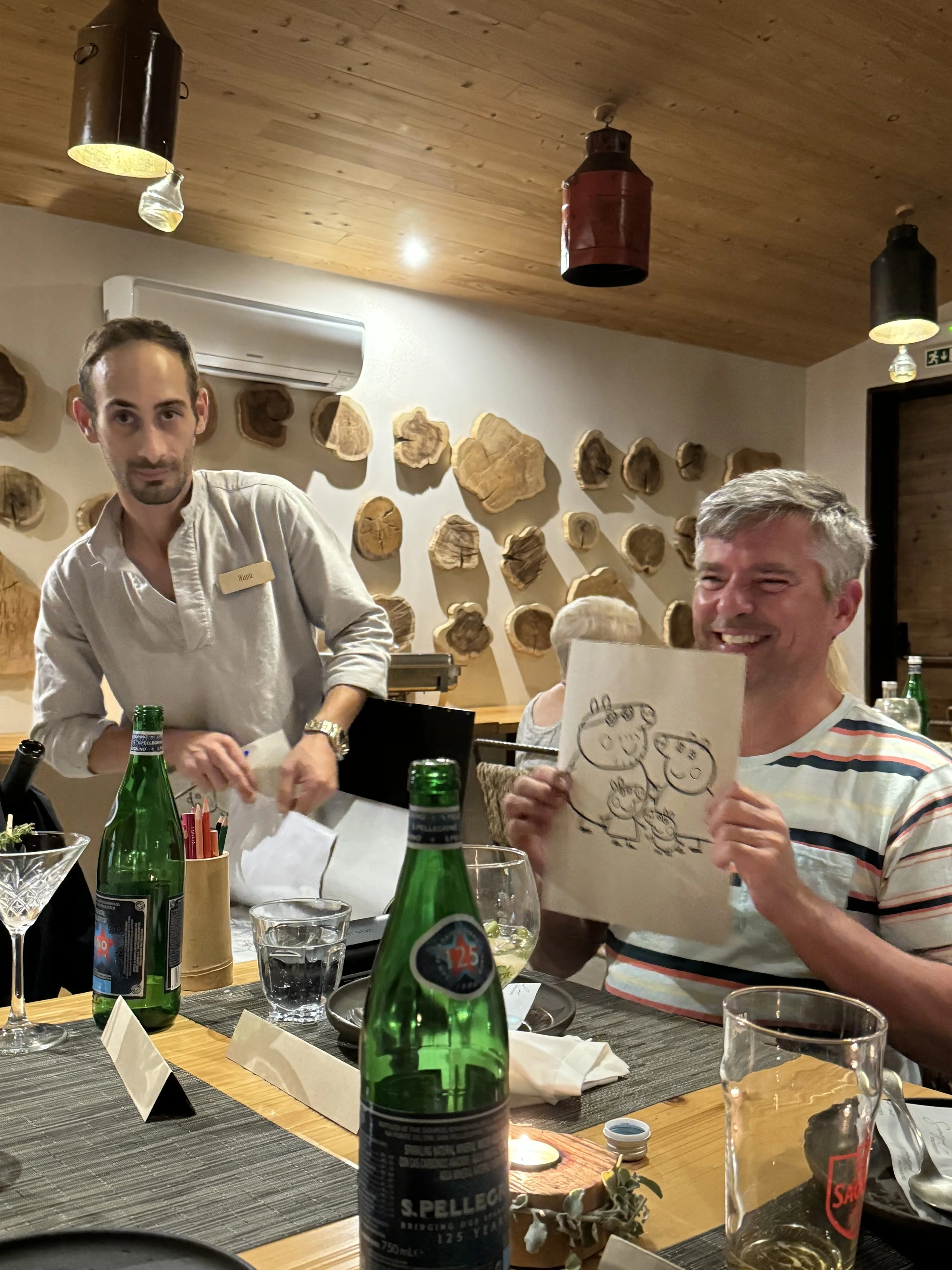 People sitting at a restaurant table enjoying a meal, with drinks, menus, and a wall decorated with wooden logs in the background.