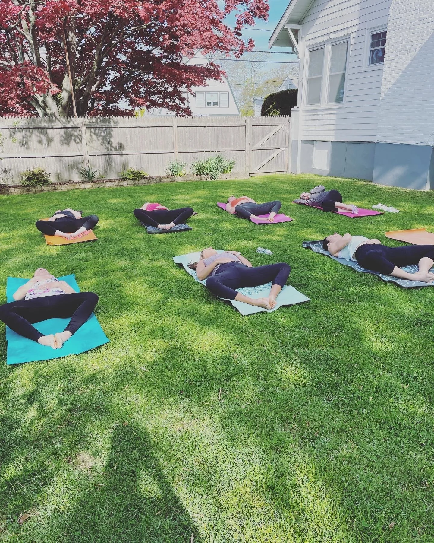 Group of people participating in outdoor yoga class on a grassy backyard, lying on colorful mats, with a white house, a fence, and trees in the background.