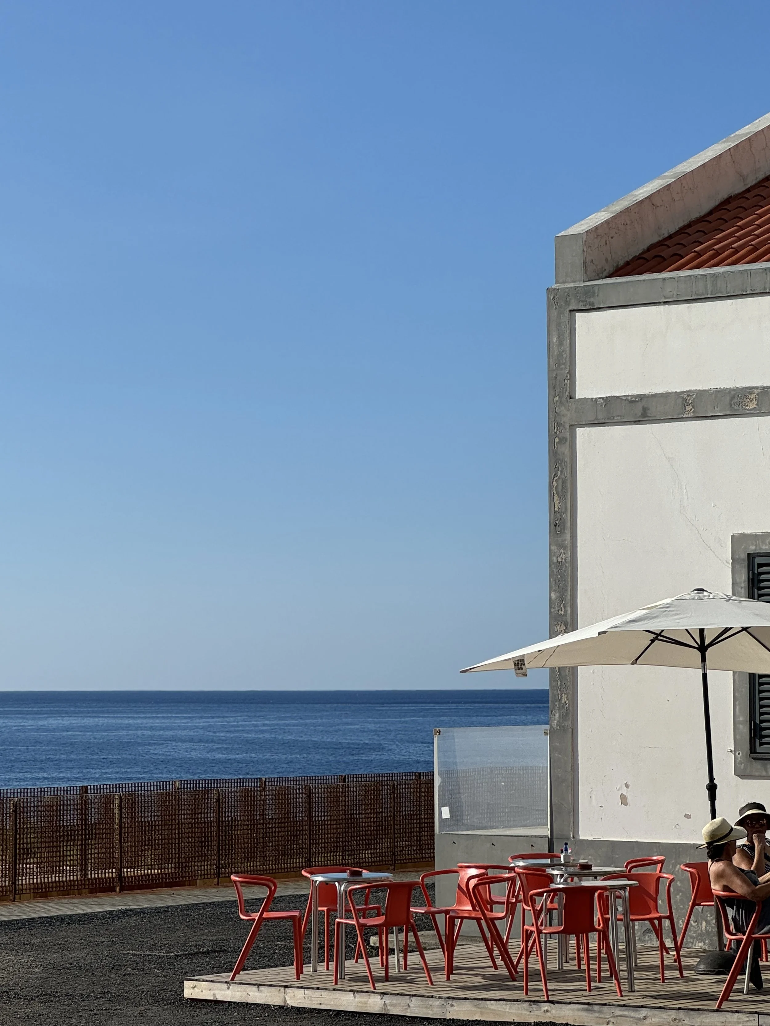 Outdoor seaside cafe with red chairs and tables under a large umbrella. A building with white walls and a sloped tiled roof is partially visible. The ocean and a clear blue sky are in the background. Two people wearing hats are seated at the table.