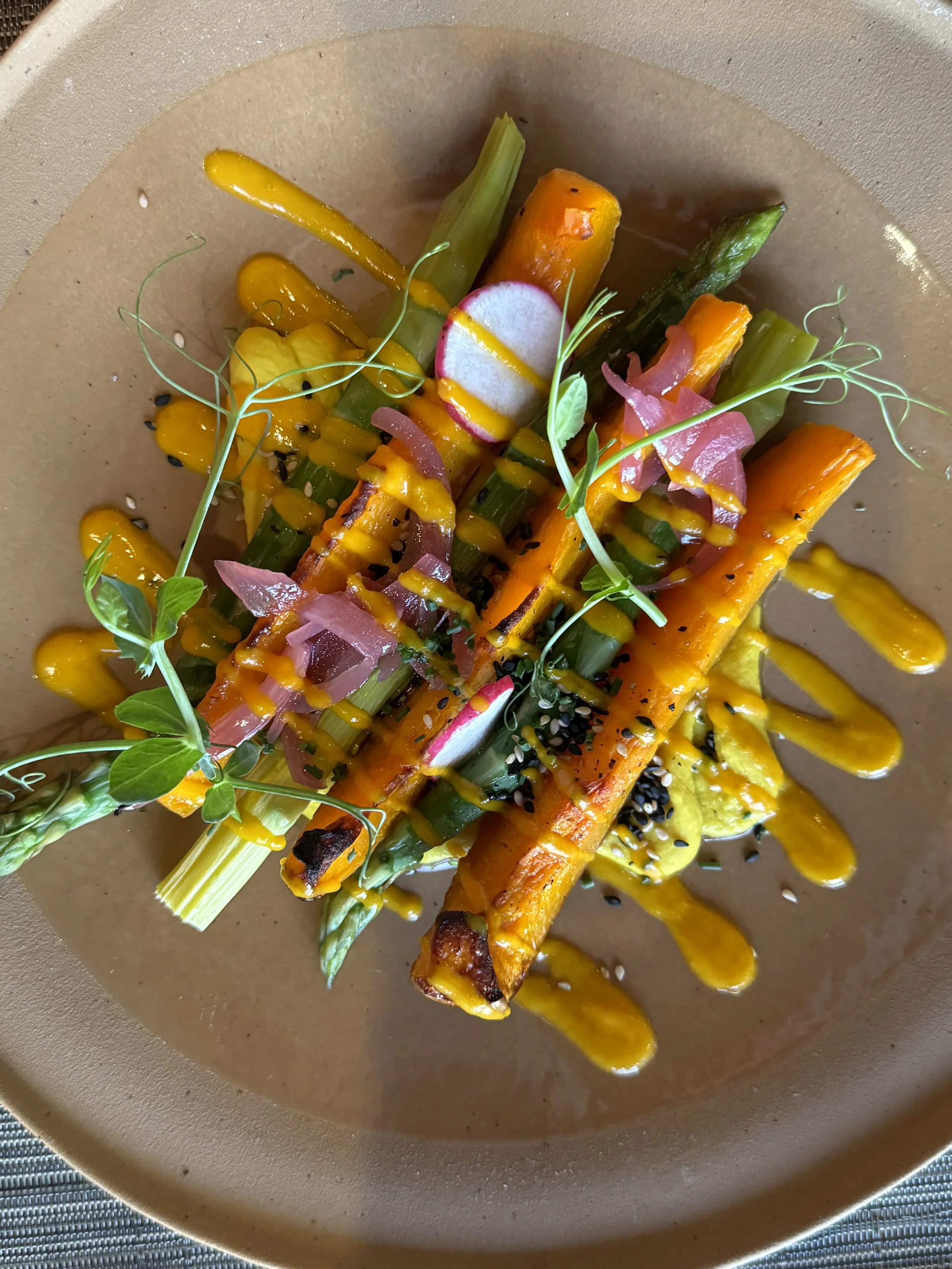 Roasted mixed vegetables on a plate with a beige background, drizzled with yellow sauce, garnished with green herbs and black sesame seeds.